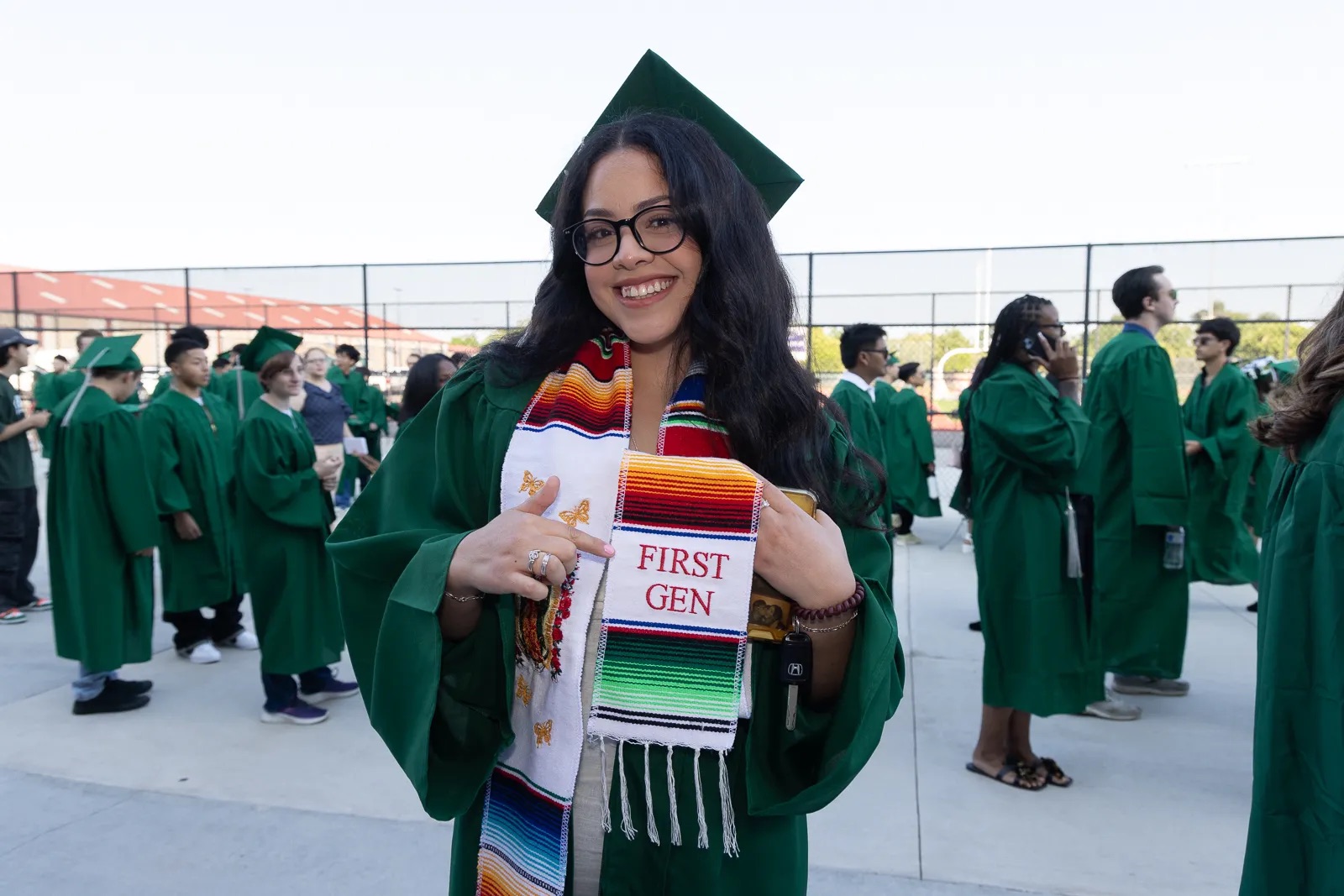 Woman in a green cap and gown holds up a stole that says first gen.