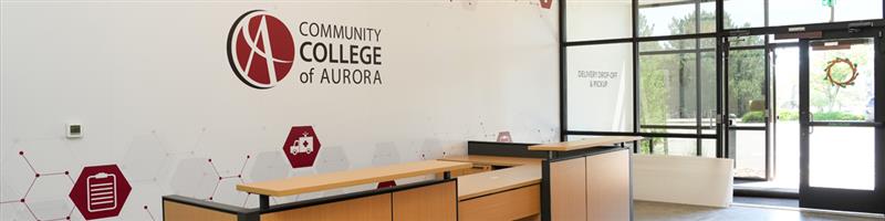 Reception area at the Community College of Aurora featuring a modern front desk and a wall with the college’s logo. The entrance doors allow natural light to enter the space, and a wreath is visible on the glass door.