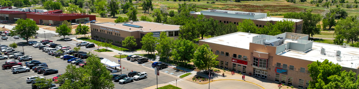 Aerial view of the Community College of Aurora’s CentreTech Campus, showing multiple academic buildings surrounded by trees, a large parking lot filled with cars, and landscaped walkways. The main entrance and signage are visible, with students walking near the front of the campus.