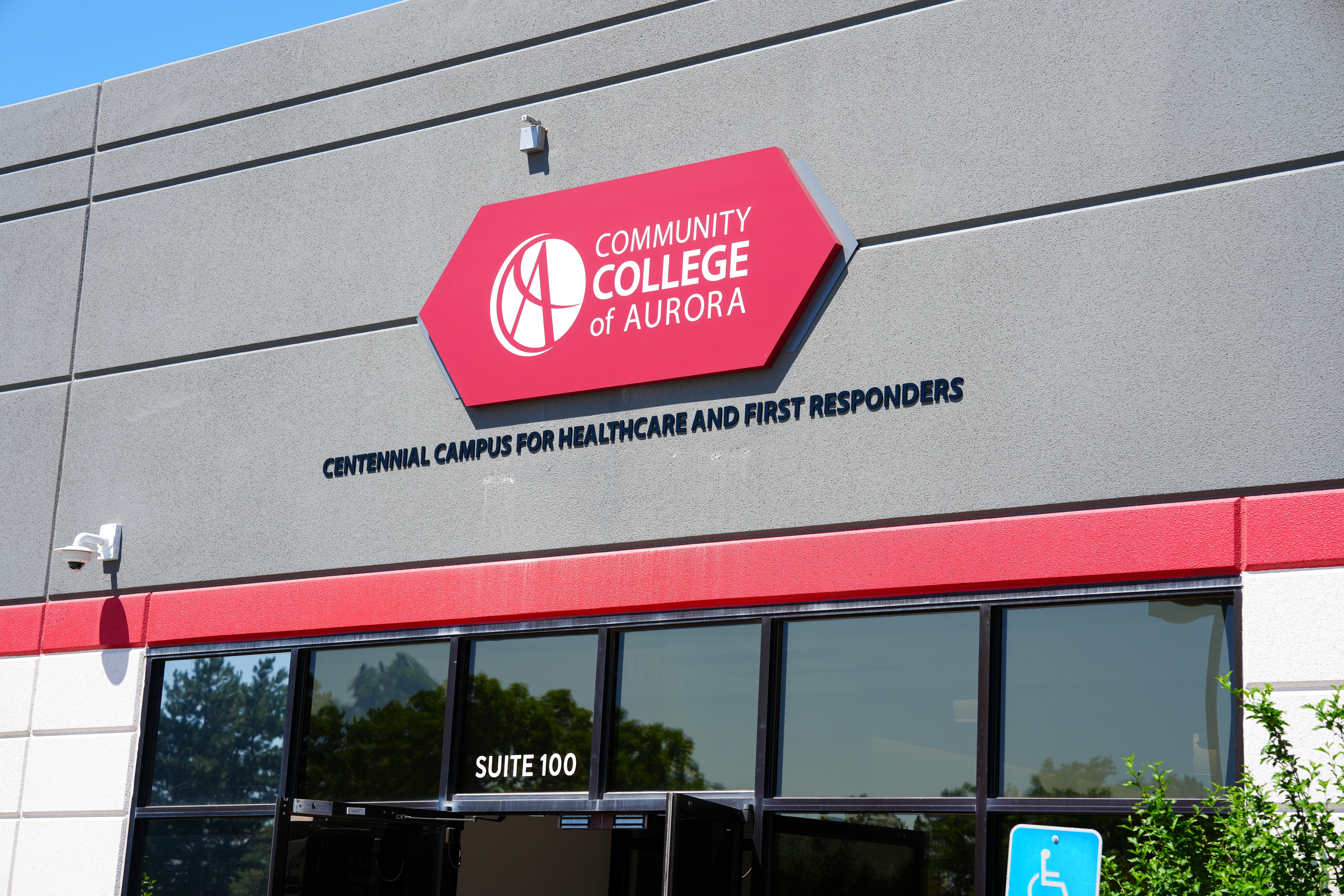 Front entrance of the Community College of Aurora’s Centennial Campus for Healthcare and First Responders. The building features a gray and red exterior with a prominent red hexagonal CCA logo and bold campus name lettering above the glass doors labeled 'Suite 100.' A blue accessible parking sign is visible in the foreground.