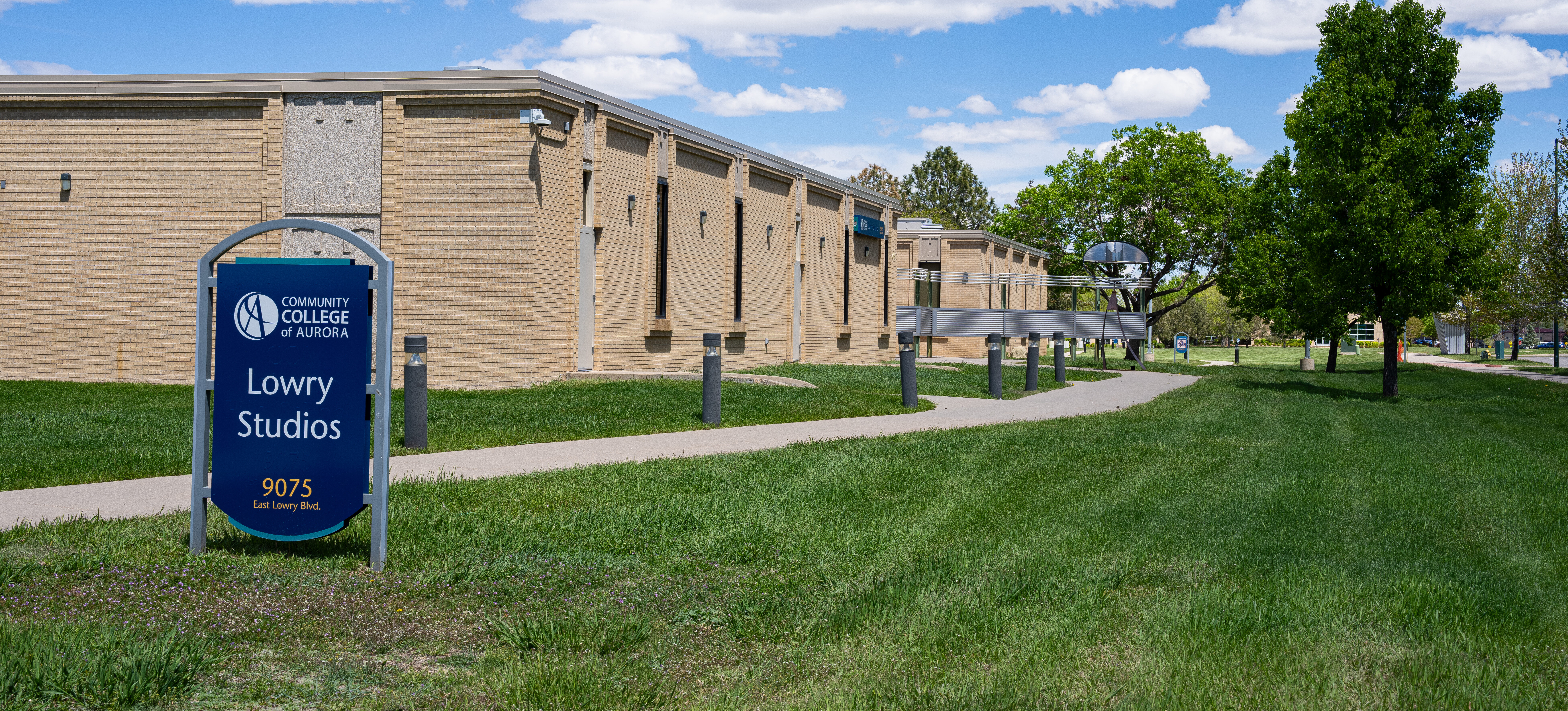 Exterior view of the Community College of Aurora’s Lowry Studios building on a sunny day, with a blue sky and scattered clouds overhead. A dark blue sign in the foreground reads “Community College of Aurora – Lowry Studios, 9075 East Lowry Blvd.” The building is a one-story brick structure, surrounded by green grass, trees, and a paved walking path.