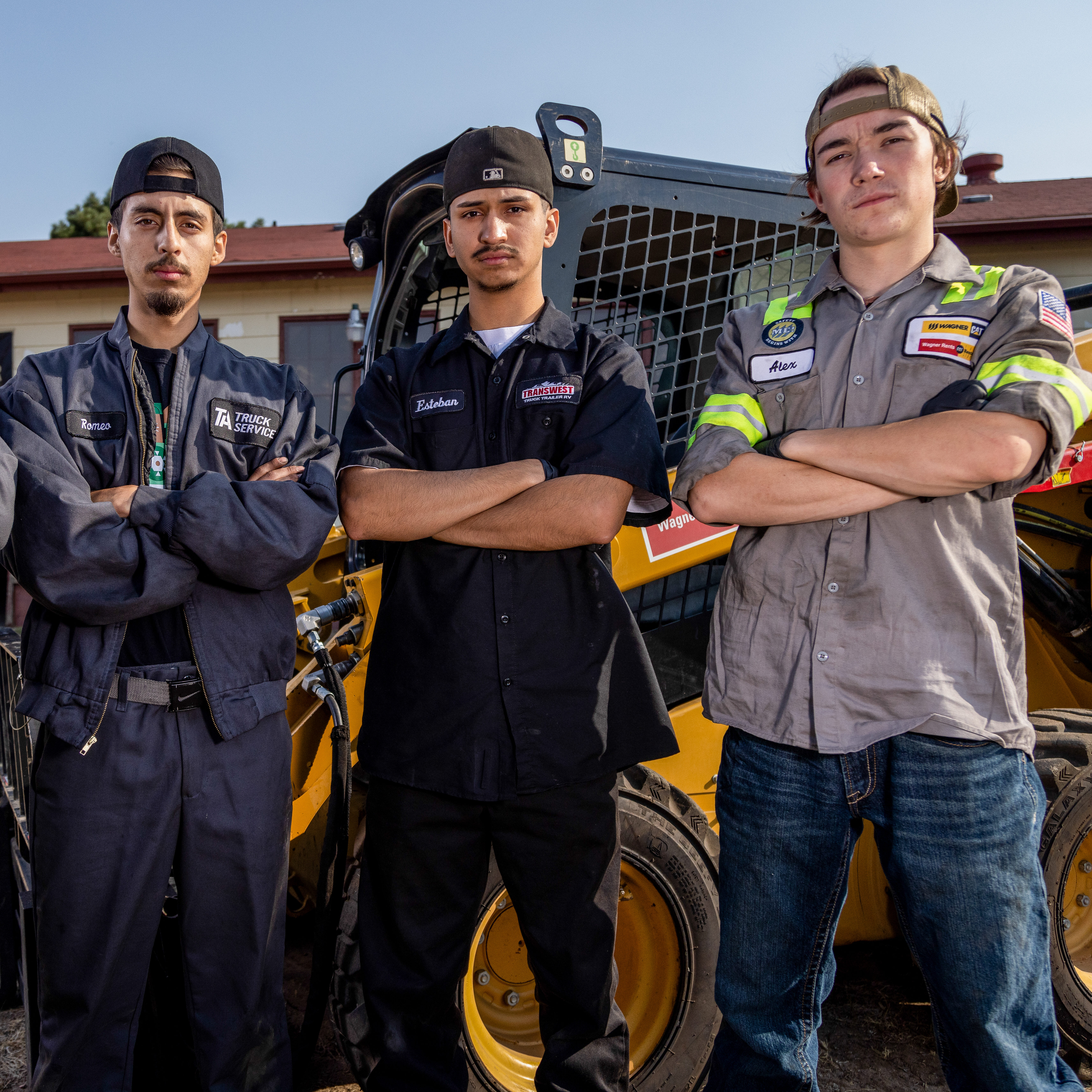 Three people stand with arms folded outdoors in front of a tractor.