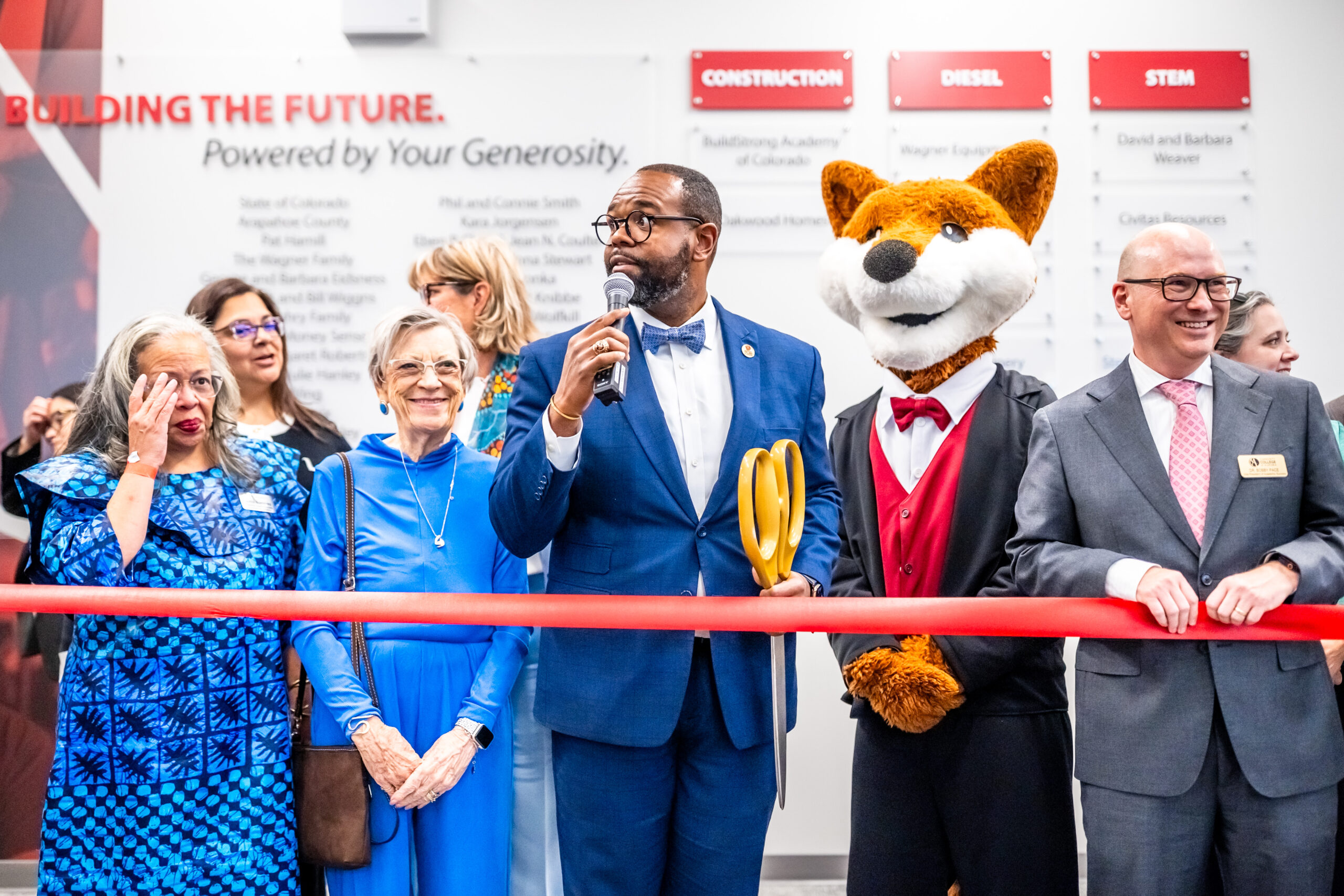 A group of people stand behind a red ribbon at a ribbon-cutting ceremony. A man in a blue suit and bow tie speaks into a microphone while holding large gold scissors. Next to him are attendees dressed formally, including a person in a fox mascot costume wearing a red vest and bow tie. A wall behind them displays donor names and headings for construction, diesel, and STEM.