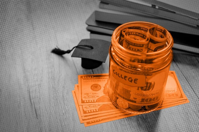 Stock photo of a jar of cash on a wooden table near a small, black graduation cap. The jar and money are tinted orange while the background is black and white.
