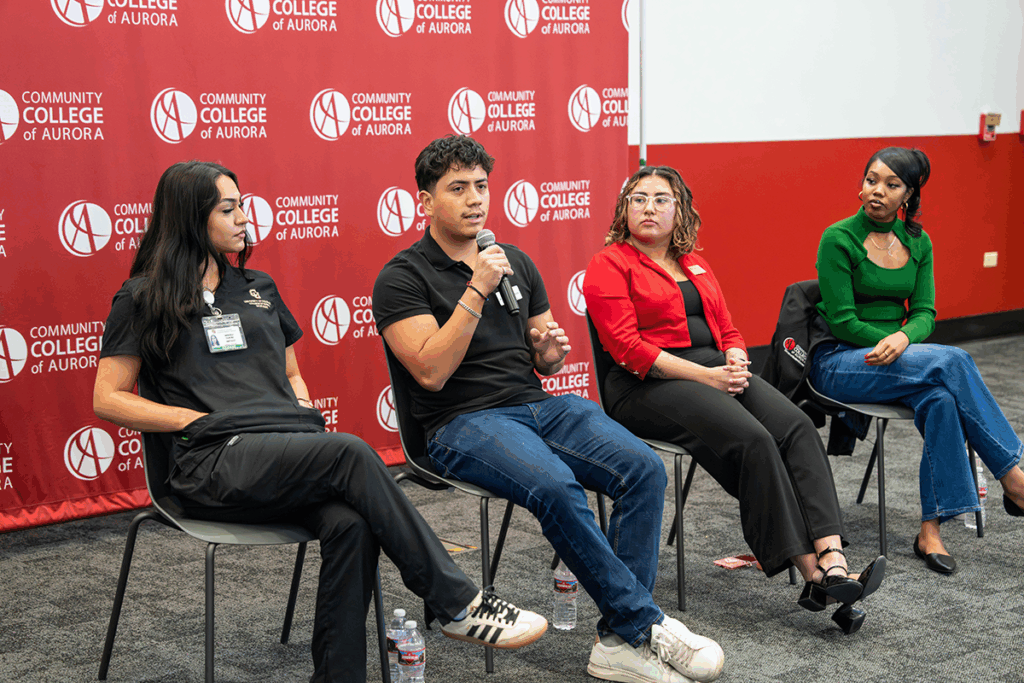 Four people sit in a row of chairs in front of a banner with the Community College of Aurora logo. One person holds and speaks into a microphone, while the others look towards them.