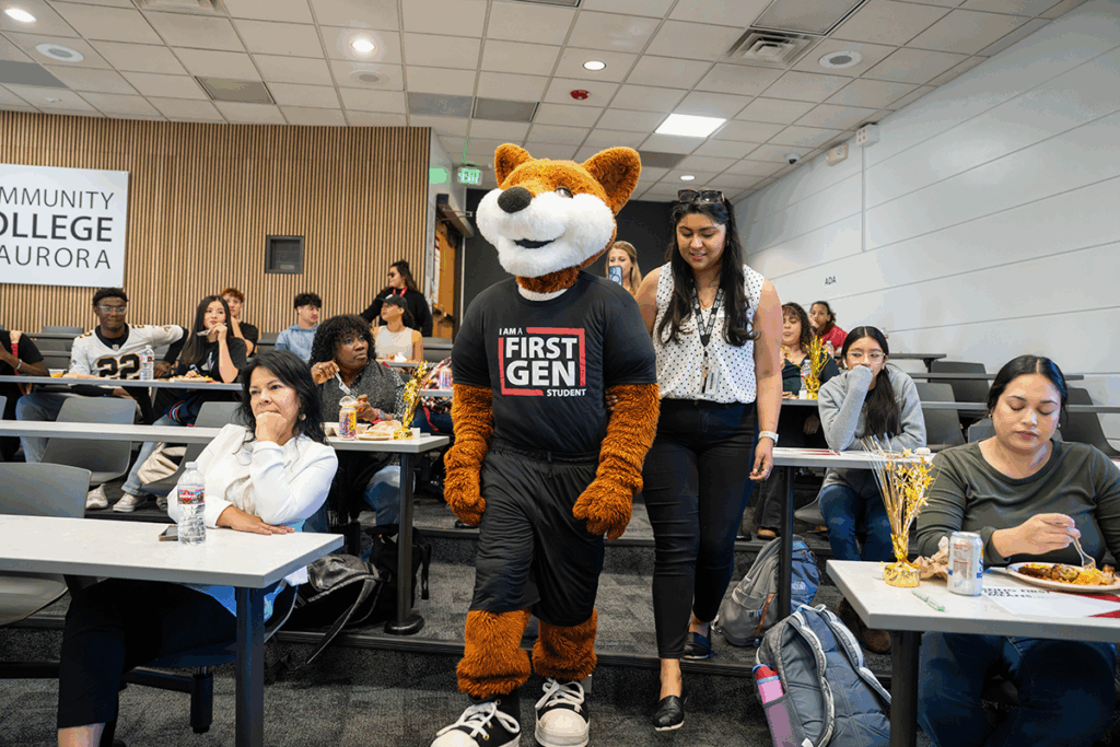 Students sit in a lecture hall while a fox mascot walks down the aisle wearing a black shirt that reads ‘I am a First Gen student.’ Some students are eating and drinking while looking at the mascot.