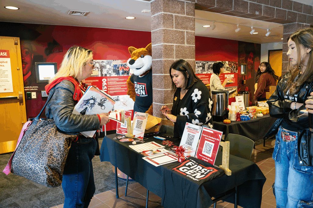 Students and staff gather at a registration table during the Community College of Aurora’s First-Generation Celebration event. The table displays flyers, QR codes, and a shirt that reads 'I am a First Gen Student.' The college mascot stands nearby, and refreshments are set up on a table in the background.