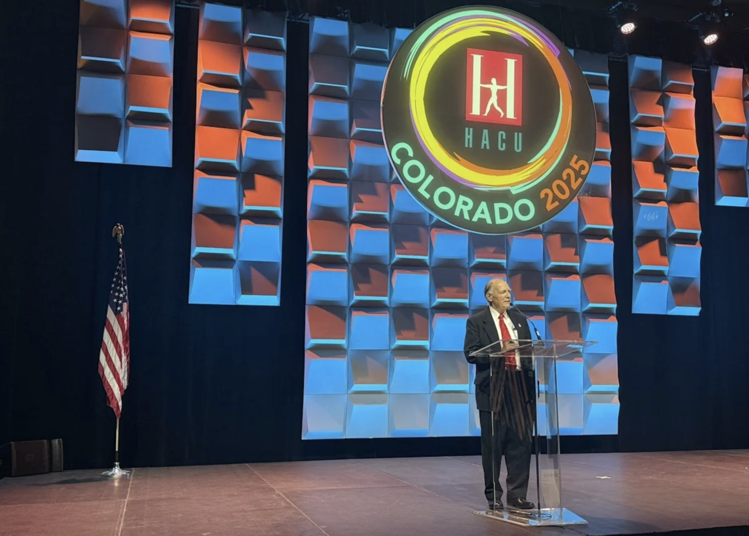A speaker stands at a clear podium on a stage with a large colorful sign reading “HACU Colorado 2025” above them. The stage backdrop features a geometric blue and orange pattern, and an American flag is positioned to the left.