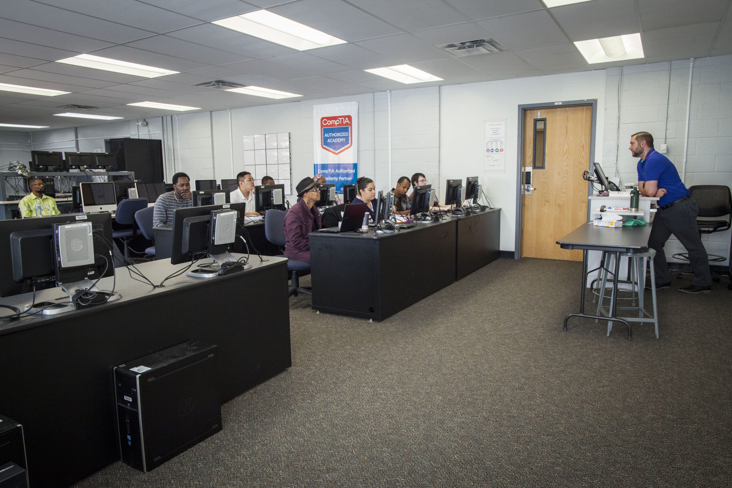 A classroom filled with students seated at computer desks, engaging in an IT training session. A teacher is standing at the front of the room, providing guidance to the students. The room contains several computers and a banner indicating it is a CompTIA Authorized Academy. The students are using computers to learn, while the teacher stands near a desk with a monitor.