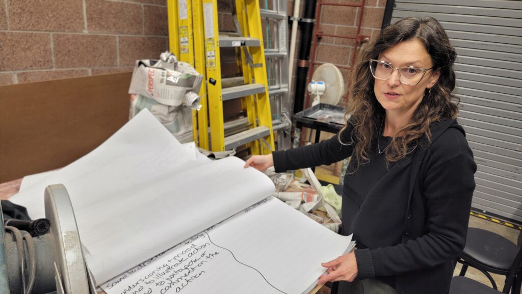 A woman wearing glasses holds open a large pad of paper covered in handwritten notes in a workshop space with ladders, tools, and supplies behind her.
