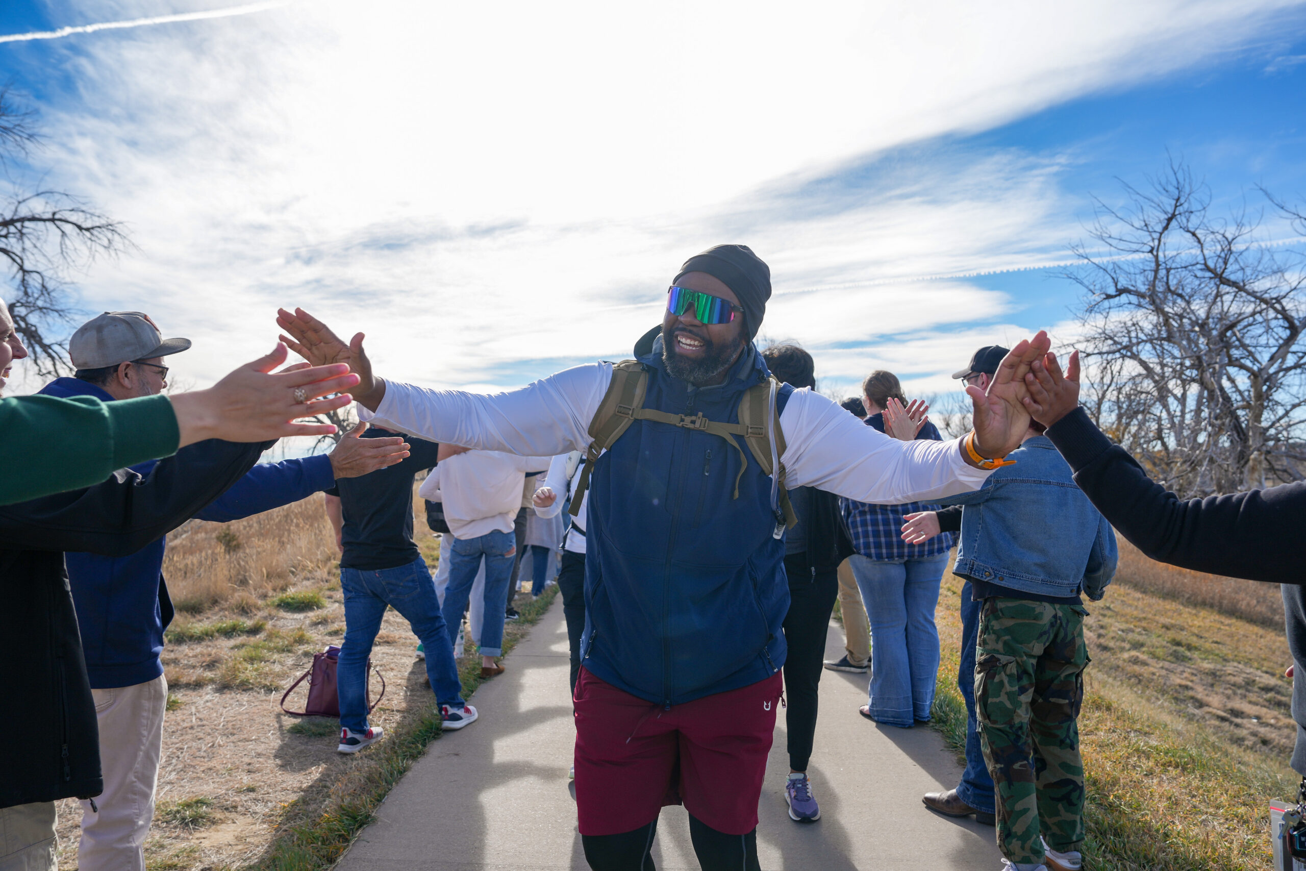 A smiling man wearing sunglasses, a beanie, and a backpack walks along an outdoor path while giving high fives to people lined up on both sides. The group stands in a sunny, open area with dry grass and leafless trees in the background.