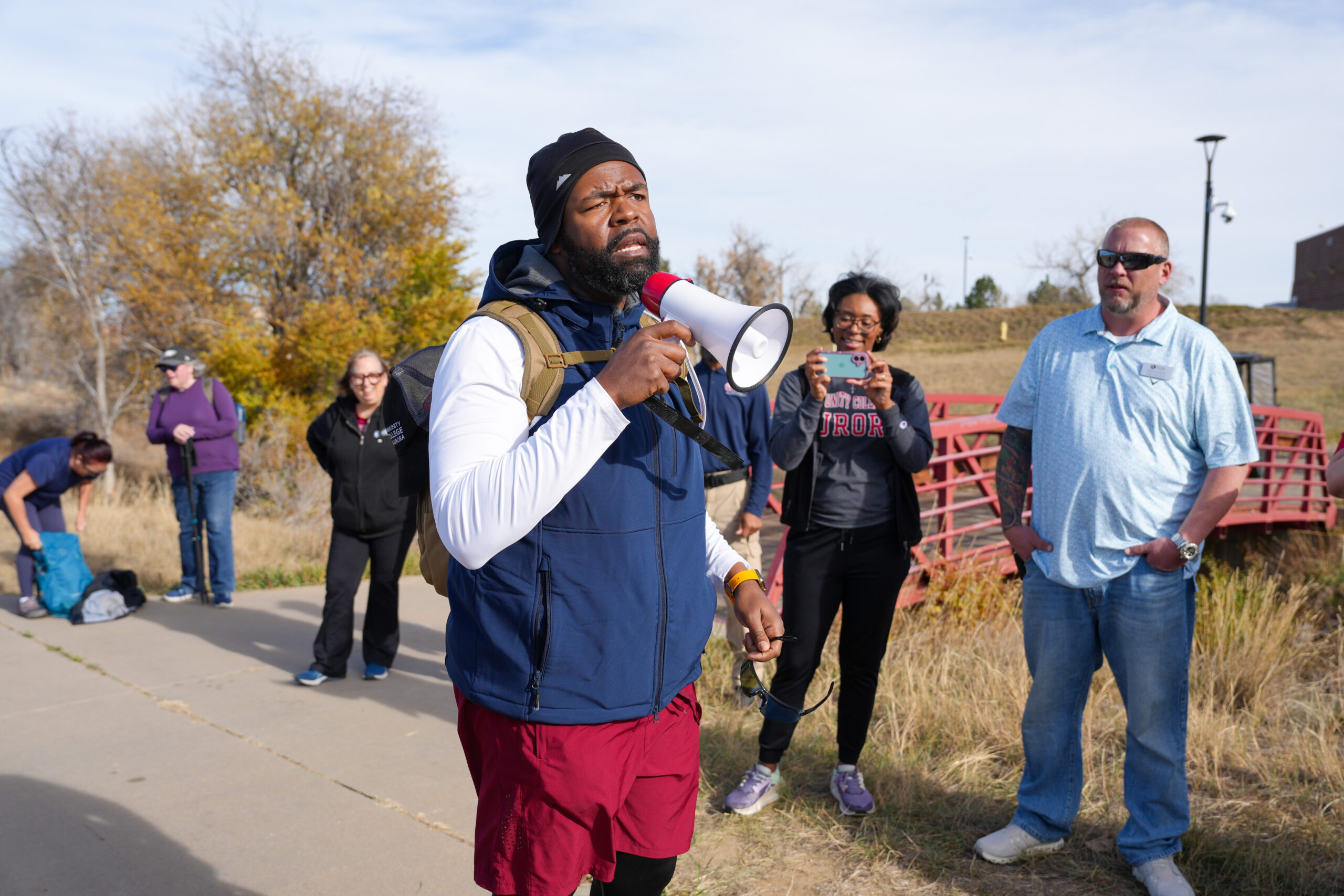 A man wearing a beanie, backpack, and athletic clothing speaks into a megaphone while standing on a paved path outdoors. Several people stand around him listening; one woman records the moment with her phone. Autumn trees and a red footbridge are visible in the background under a partly cloudy sky.