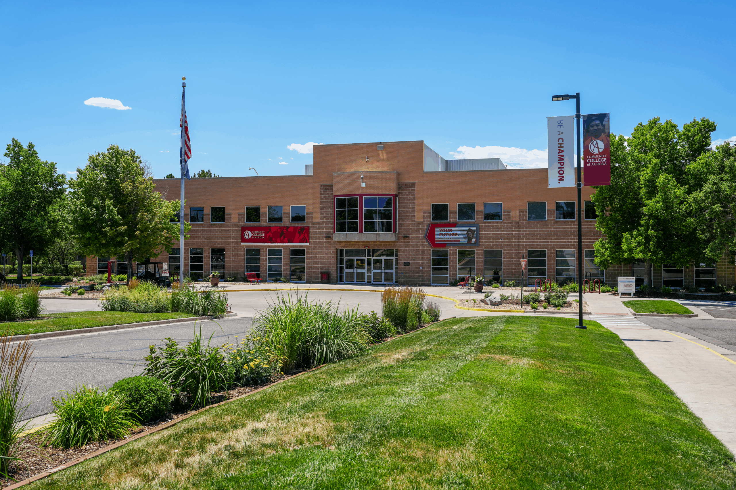Front view of the CentreTech campus main building at the Community College of Aurora on a sunny day, showing a brick building with large windows, landscaped greenery, an American flag, and campus banners reading 'Be a Champion' and 'Your Future'.