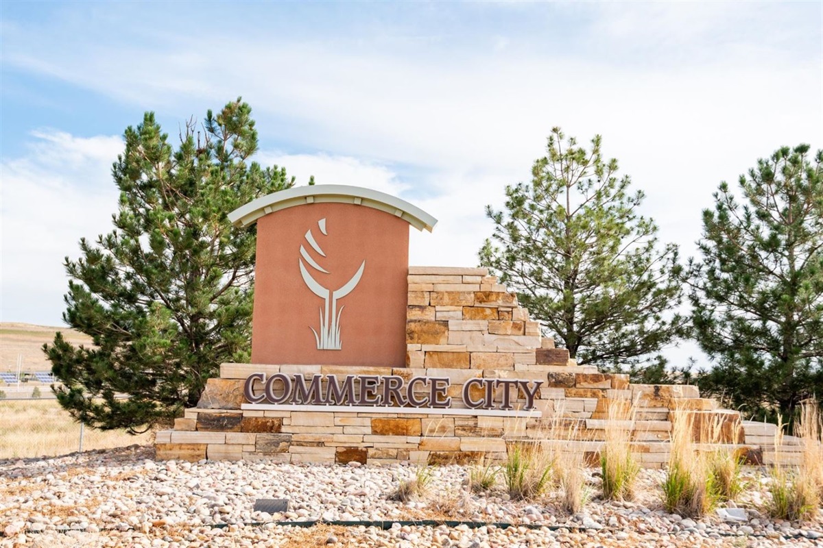A stone monument sign reading ‘Commerce City’ stands in front of pine trees. The sign features layered tan and brown stonework with a curved metal top and a stylized leaf logo on an orange panel. Rocks and dry grasses surround the base under a bright, partly cloudy sky.