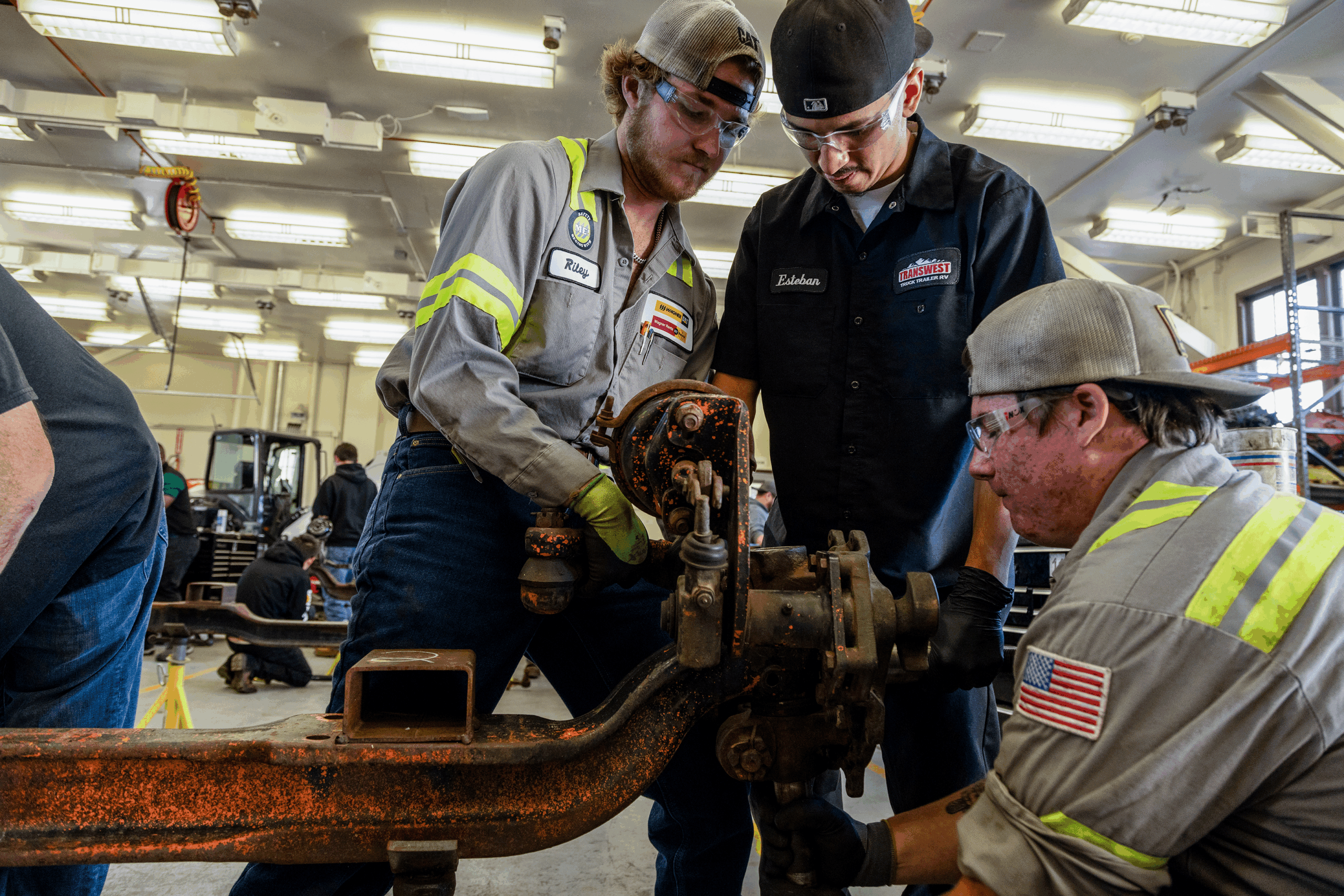 Three mechanical workers in a workshop circled around an automotive vehicle axle and using tools to work on it.