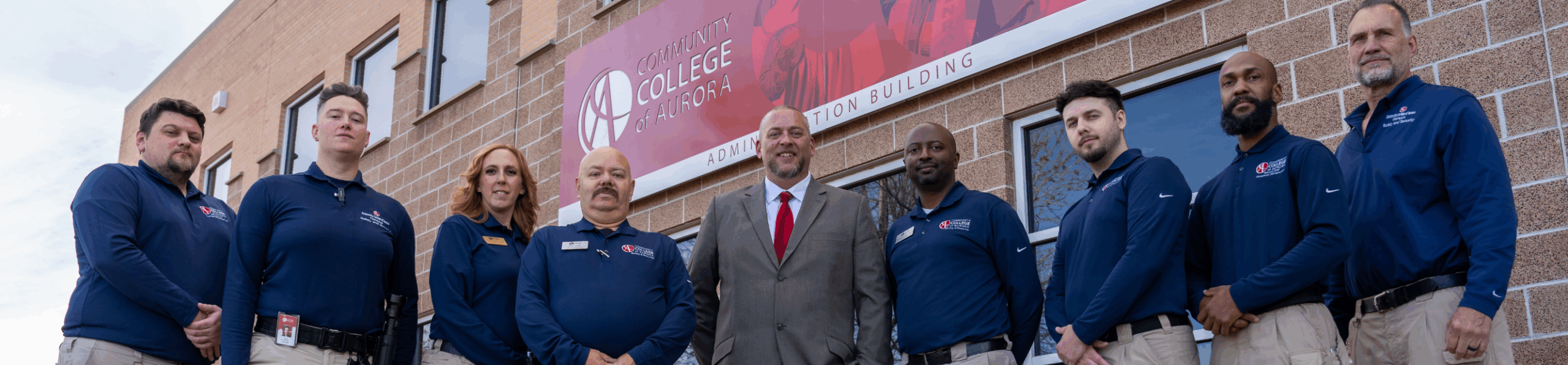 A group of ten Community College of Aurora campus safety staff members stand in a line outside the Administration Building. Most are wearing navy blue CCA security uniforms with khaki pants, while one person in the center wears a gray suit with a red tie. They pose in front of a brick building with a large CCA sign above them.