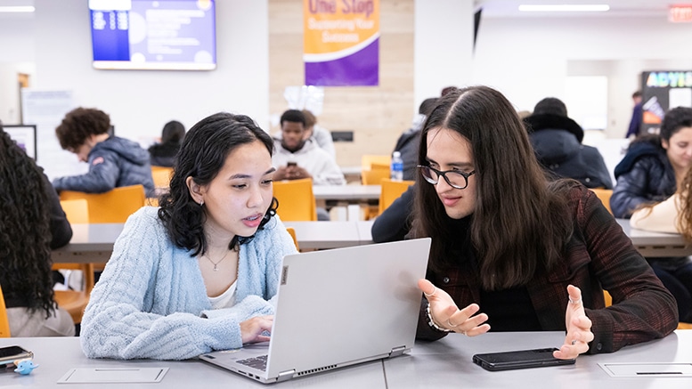 Two students sit together at a table in a busy campus study area, looking at a laptop. One student types while the other gestures as they talk through something on the screen. Other students work in the background near a sign that reads “One Stop Supporting Your Success.”
