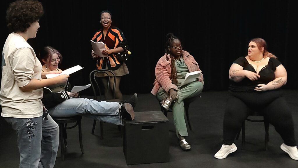 A group of five students sit and stand in a black box theater, smiling and holding scripts during a rehearsal.