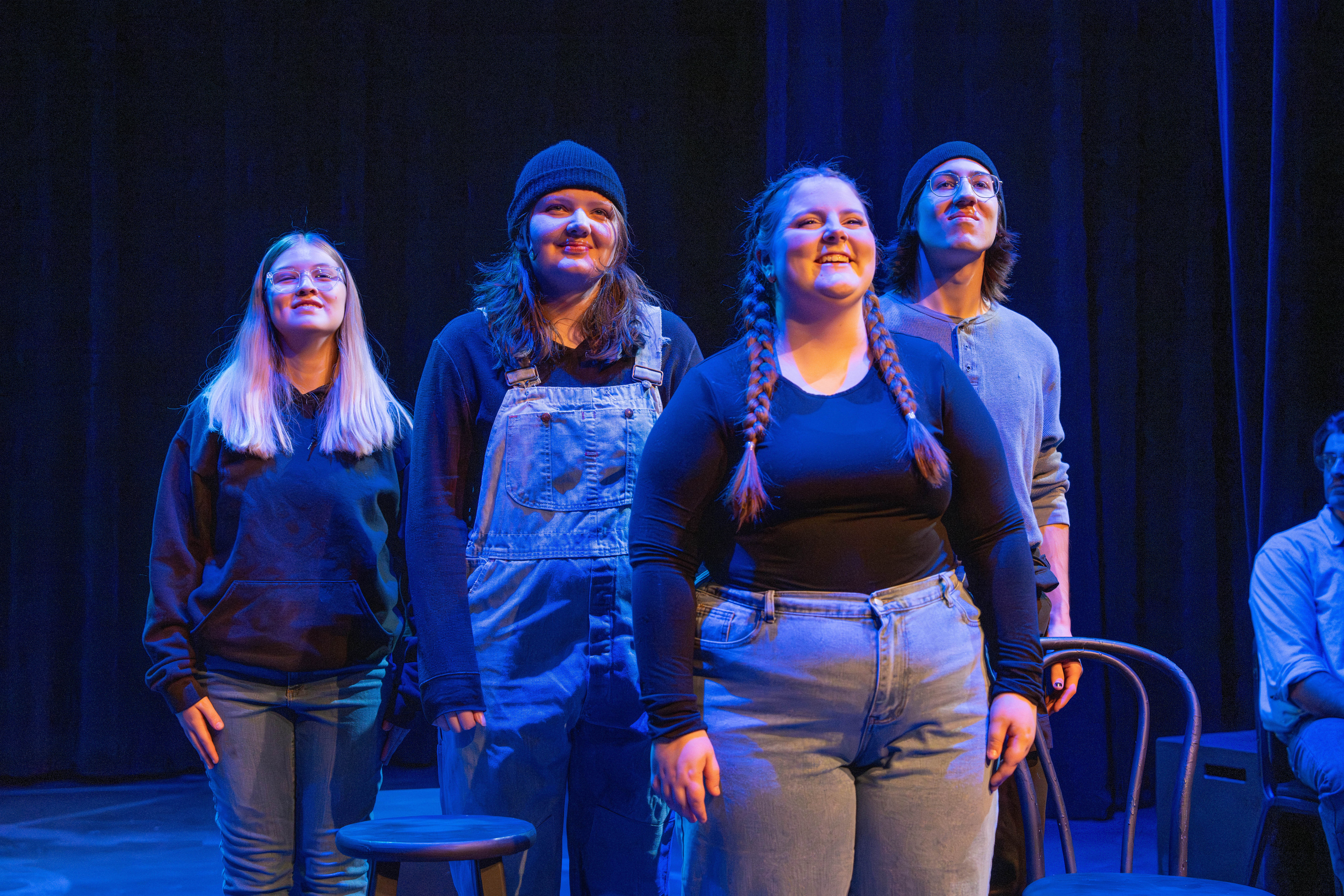 Four theatre students stand on stage under blue and purple lighting during a performance or rehearsal. They face forward with confident expressions, dressed in casual clothing including hoodies, overalls, and jeans. Empty chairs and stools are visible around them, with a dark curtain backdrop behind.