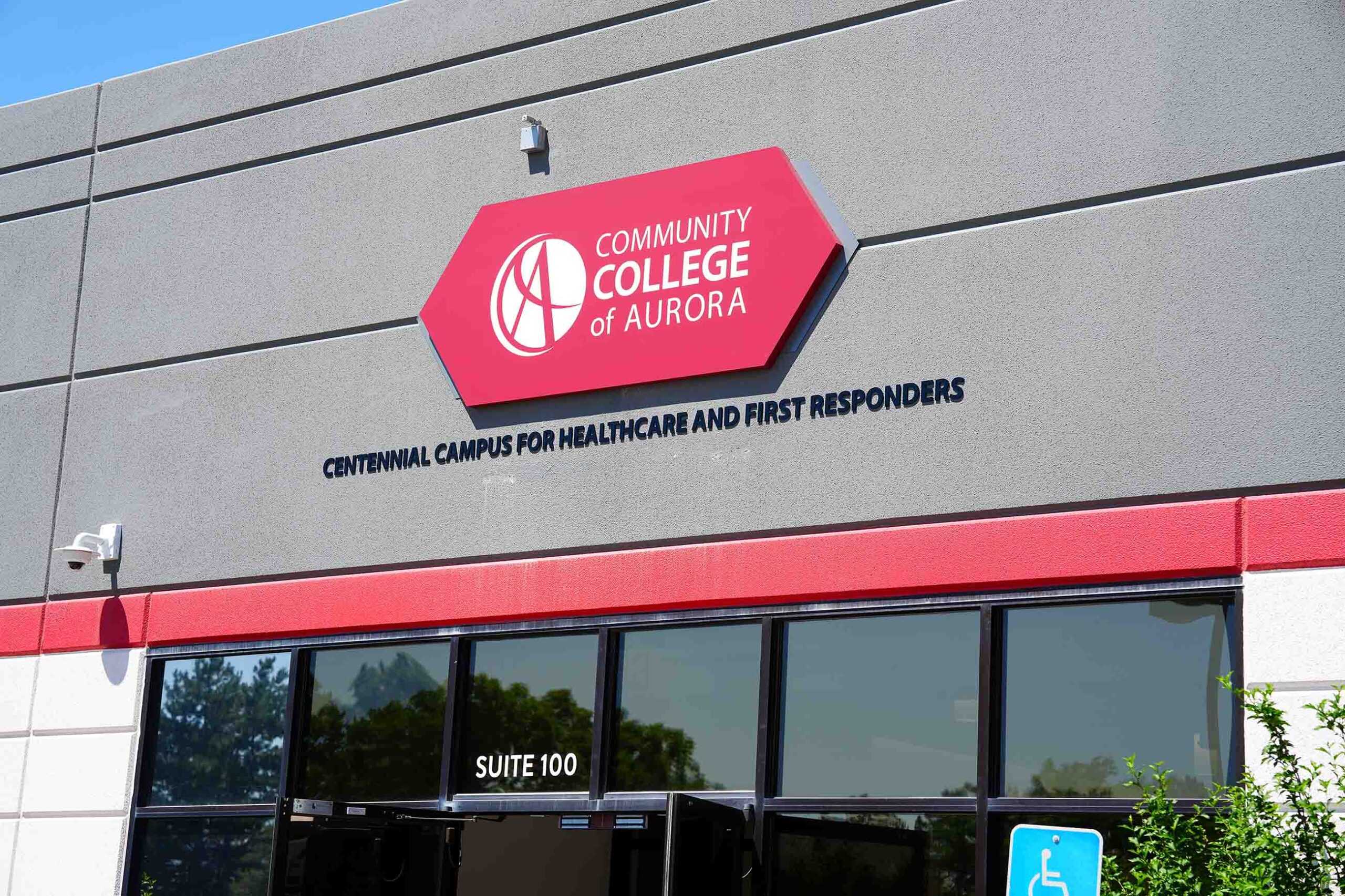 Front entrance of the Community College of Aurora’s Centennial Campus for Healthcare and First Responders. The building features a gray and red exterior with a prominent red hexagonal CCA logo and bold campus name lettering above the glass doors labeled 'Suite 100.' A blue accessible parking sign is visible in the foreground.