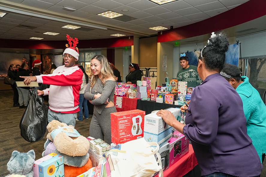 People browse tables filled with toys, games, and gift items at an indoor holiday shop, with several attendees wearing festive clothing and interacting with vendors.