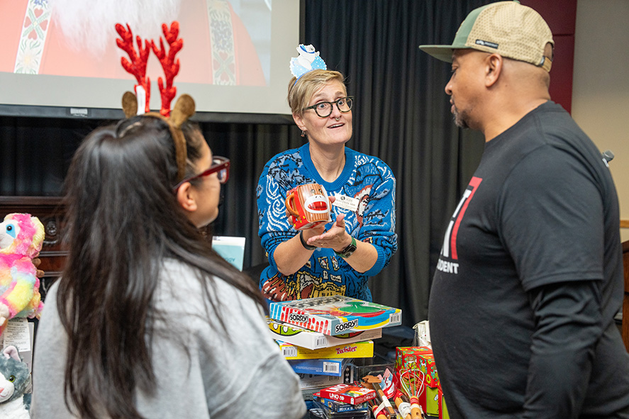 Three people stand around a table of toys and games at a holiday event while a person in a festive sweater and headband holds up a toy and talks with them.
