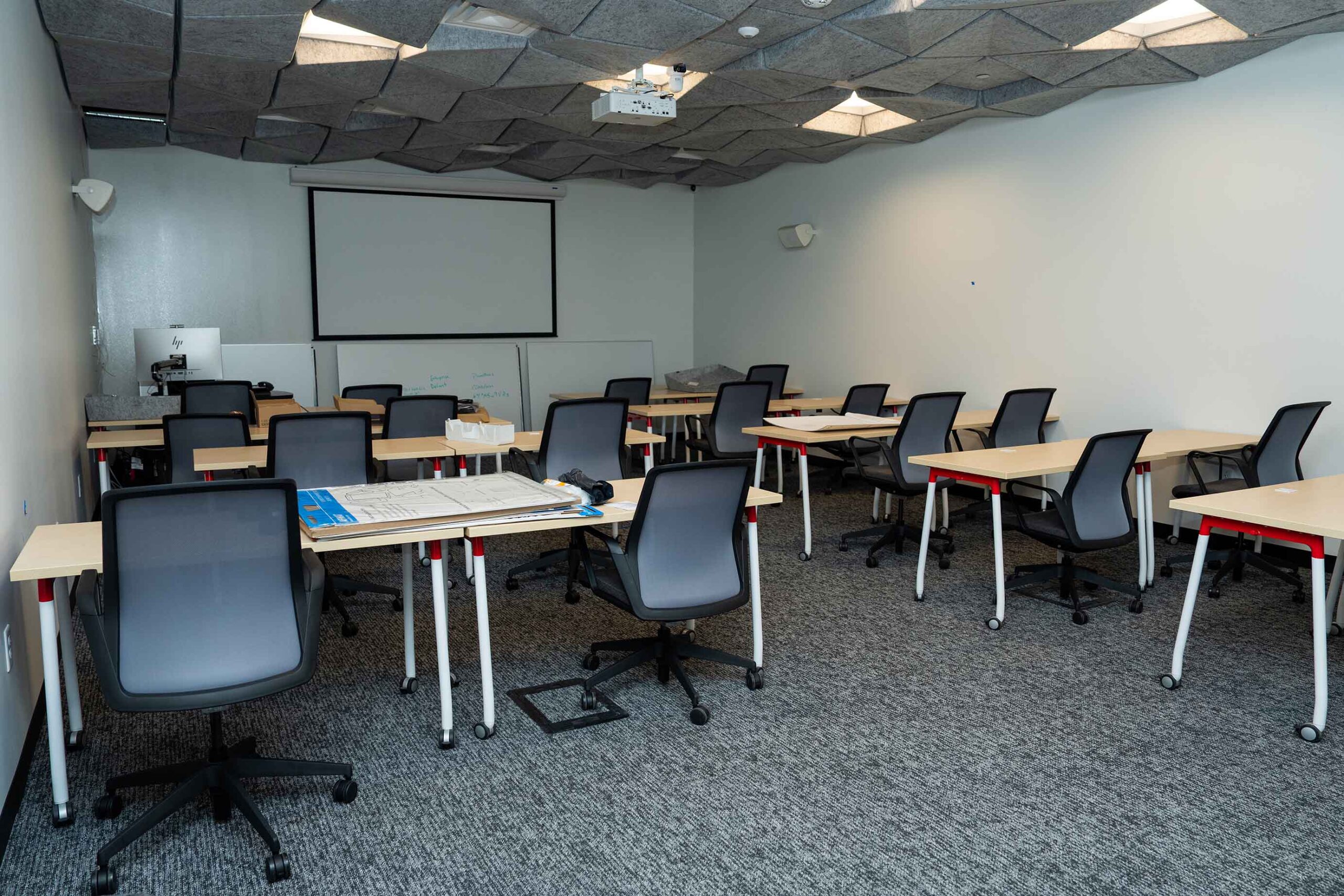 A modern classroom with multiple desks and office chairs arranged in rows. A blank projector screen is on the wall, exuding a professional ambiance.