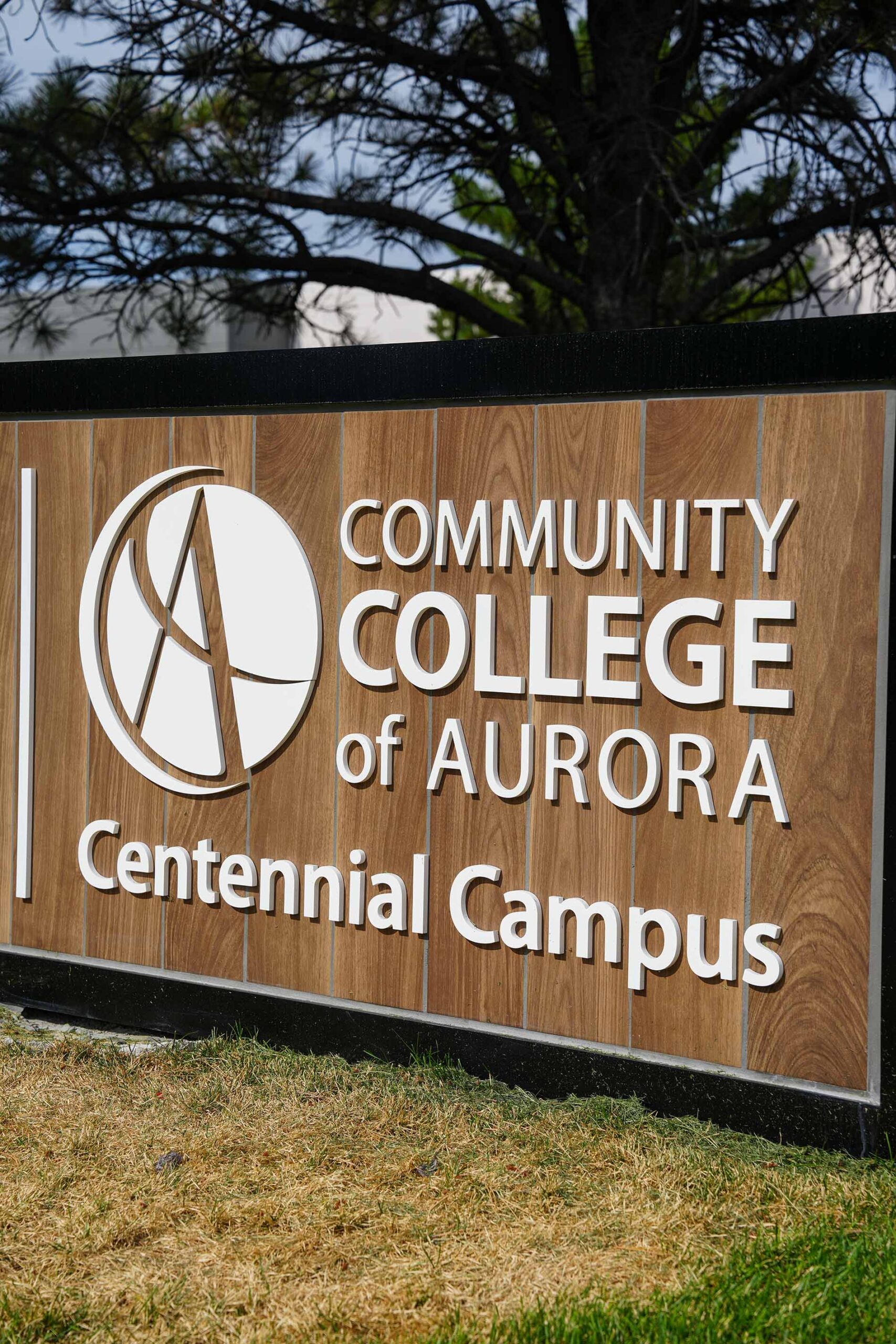 Sign for Community College of Aurora, Centennial Campus, displayed on a wooden board with white text. Background features green grass and a tree.