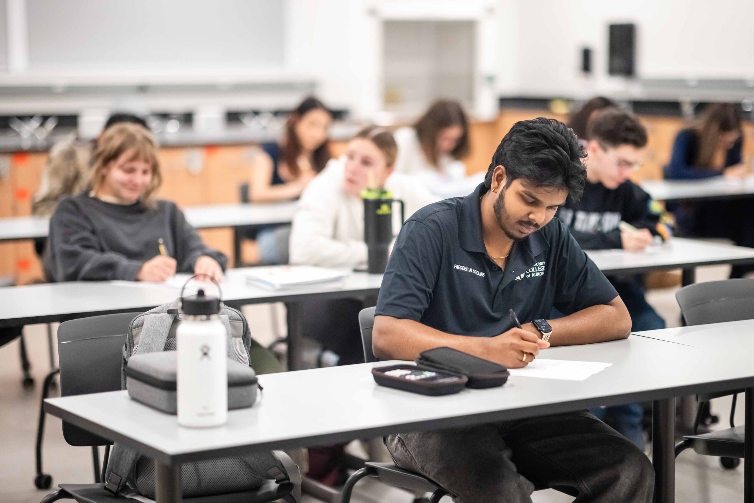 A classroom scene with diverse students sitting at desks, focused on writing. A student in a dark shirt is in the foreground, conveying concentration.