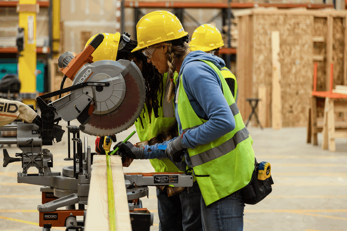 Two people wearing yellow hard hats, safety glasses, and high-visibility vests work together in a construction lab, measuring a long piece of lumber next to a table saw. Shelving and partially built wooden structures are visible in the background.