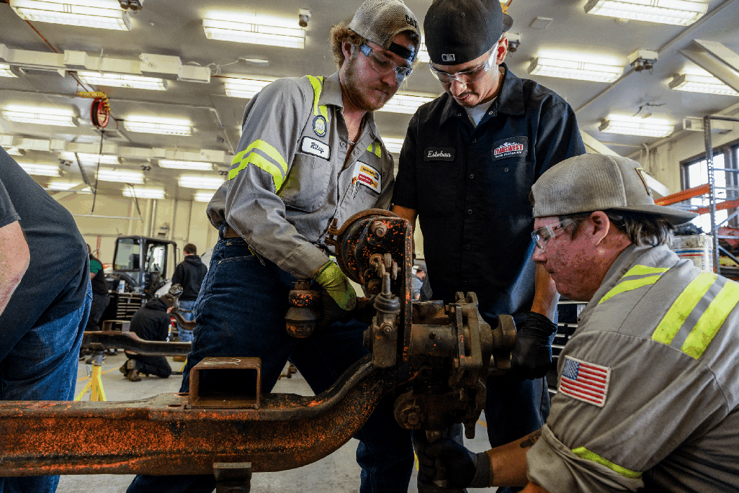 Three mechanical workers in a workshop circled around an automotive vehicle axle and using tools to work on it.