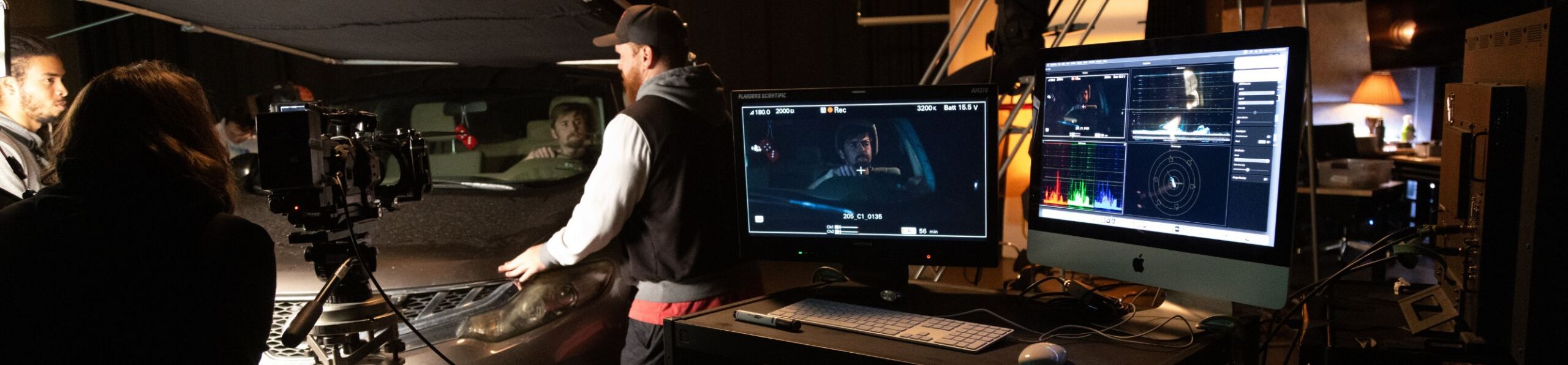 Film crew on a studio set records a scene with a car, using a mounted camera and lighting rig, while two computer monitors display the live camera feed and color correction tools.
