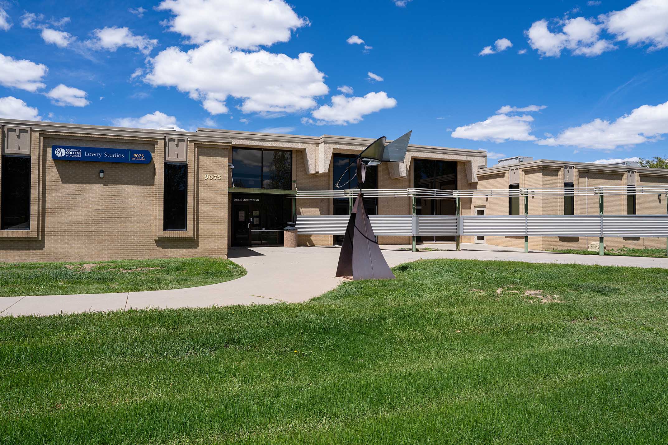 Exterior view of the Community College of Aurora's Lowry Studios building, located at 9075 East Lowry Boulevard. The building features tan brick architecture, large windows, and a blue sign displaying the college logo. A modern metal sculpture stands in the foreground along a curved sidewalk, with a bright blue sky and scattered clouds overhead.