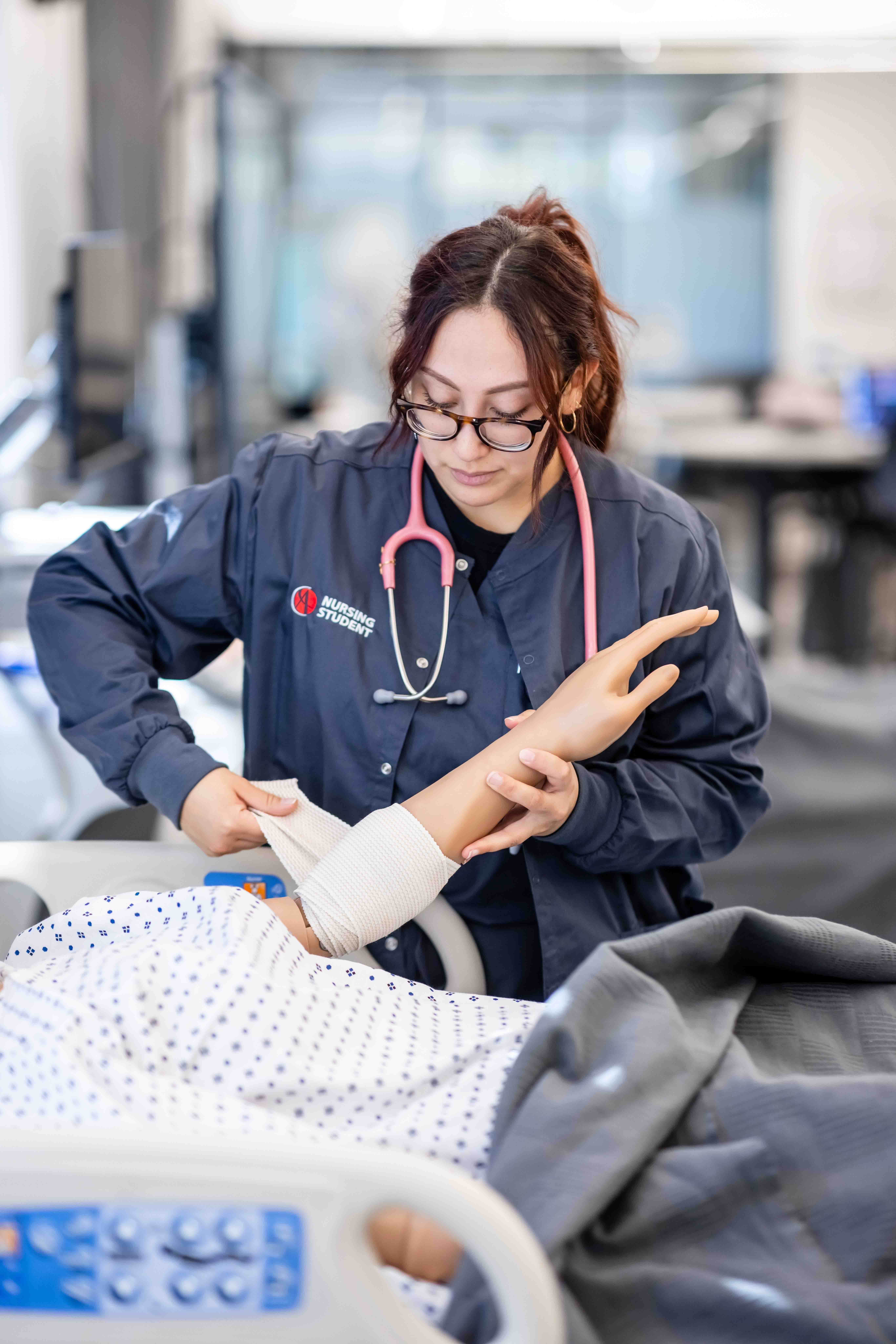 A nursing student with glasses, wearing a stethoscope, practices bandaging on a mannequin's arm in a clinical setting, conveying focus and learning.