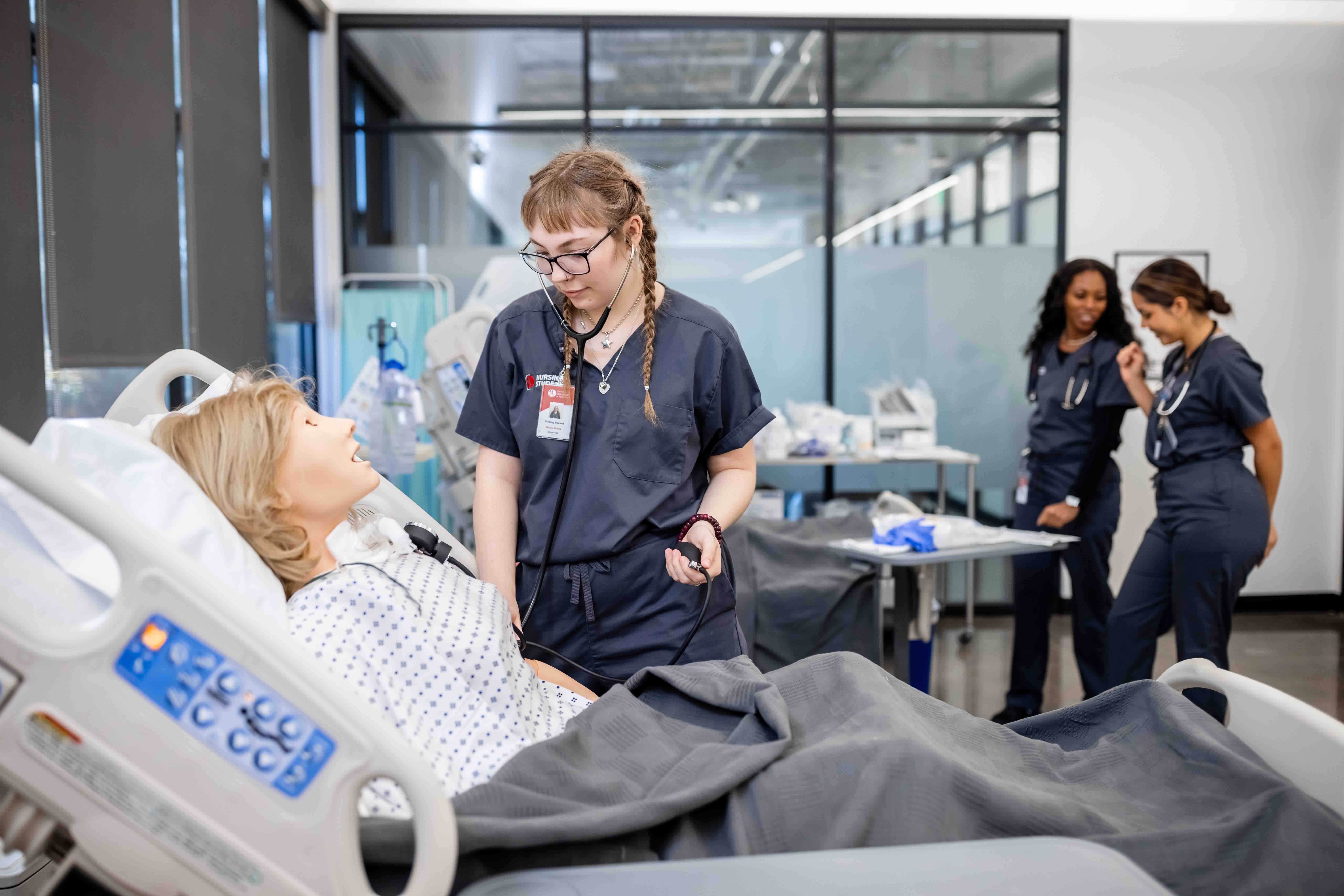 A nurse is attending to a patient mannequin in a hospital-like training setting. The mannequin, lying in a hospital bed, is dressed in a patient gown, while the nurse, wearing scrubs, listens to the mannequin's chest with a stethoscope. In the background, other healthcare workers in scrubs are standing near medical equipment and supplies.
