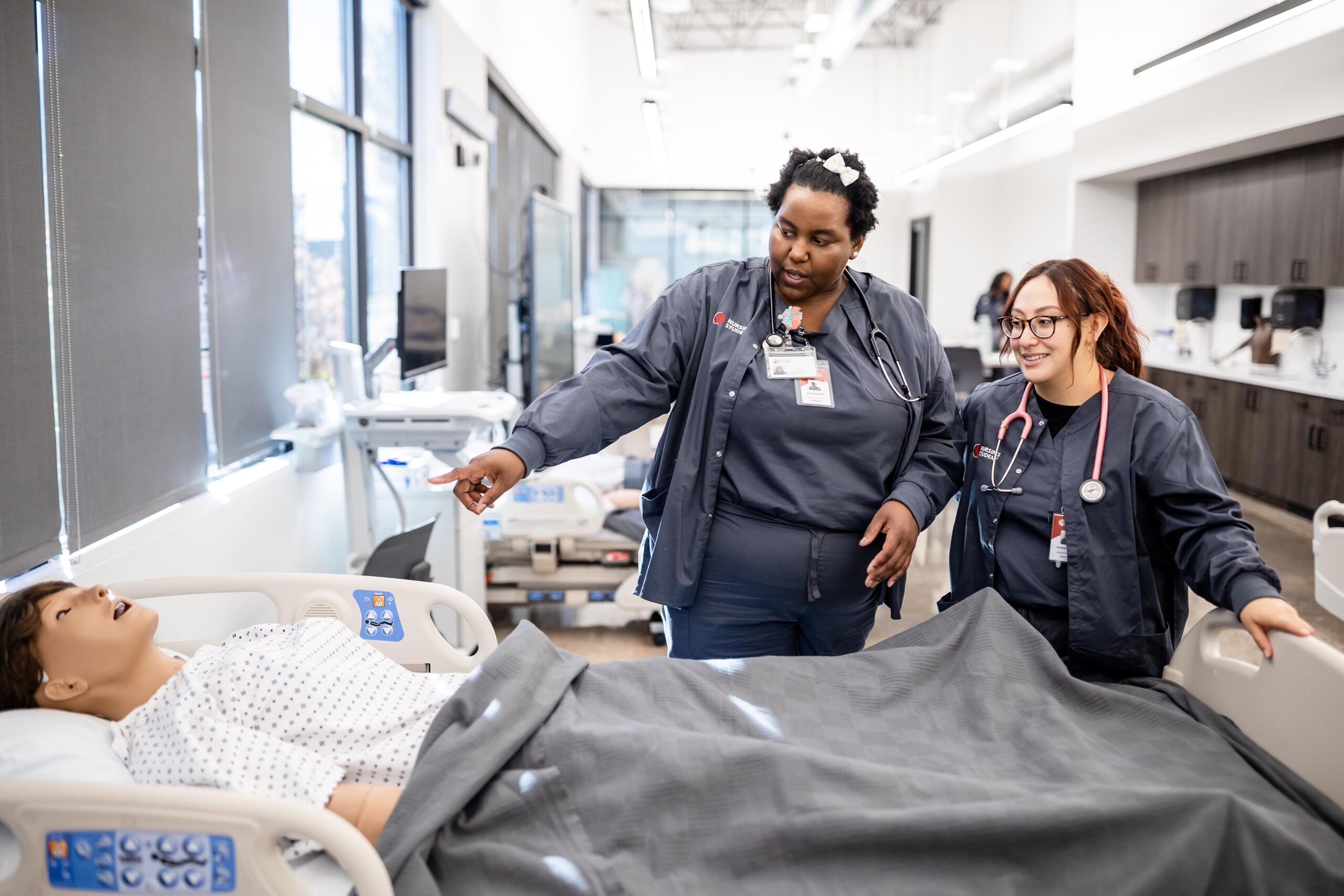 Two healthcare workers are in a medical training room, engaged with a patient simulator on a hospital bed. They seem focused and collaborative.