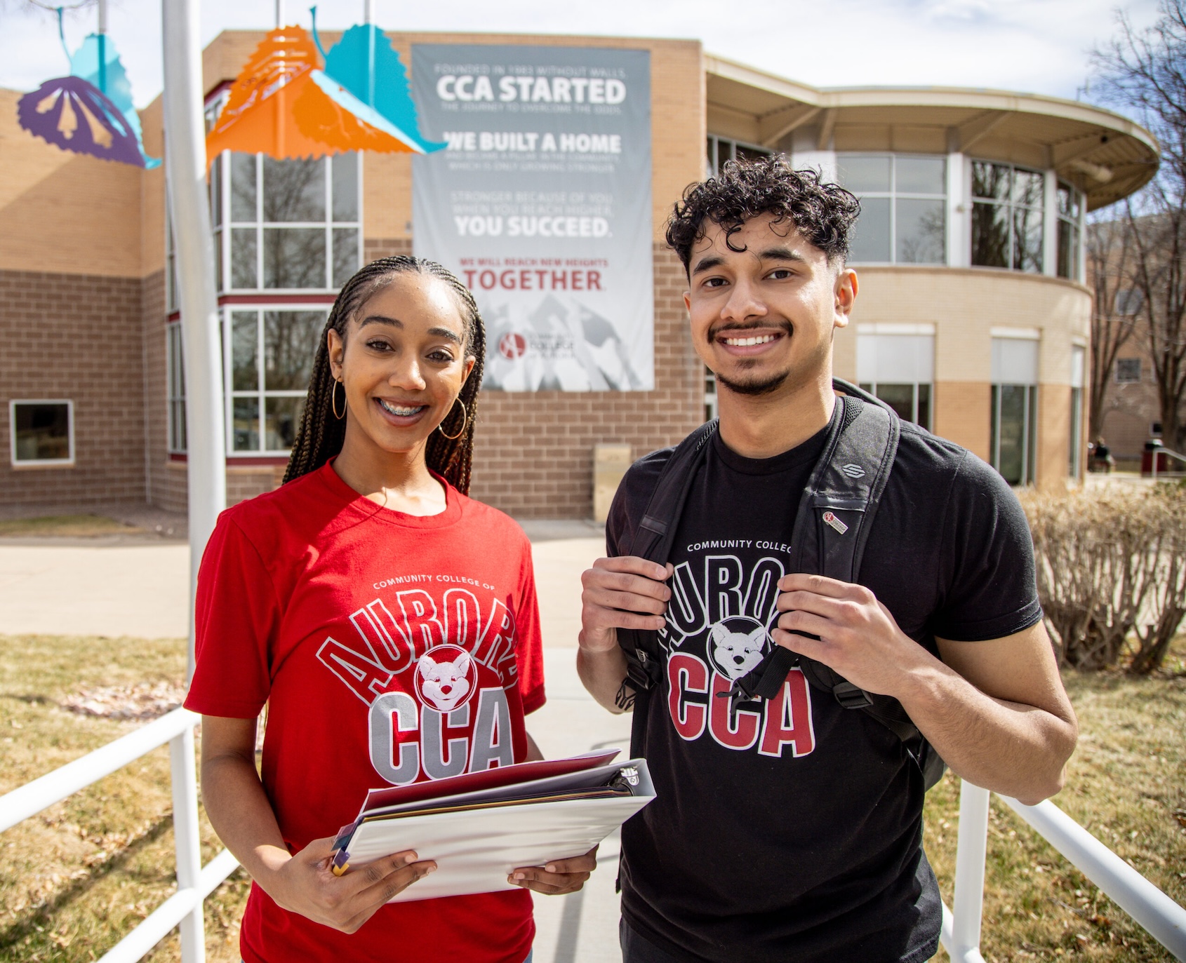 Two smiling people standing outdoors wearing shirts for the Community College of Aurora. A building stands behind them in the background.