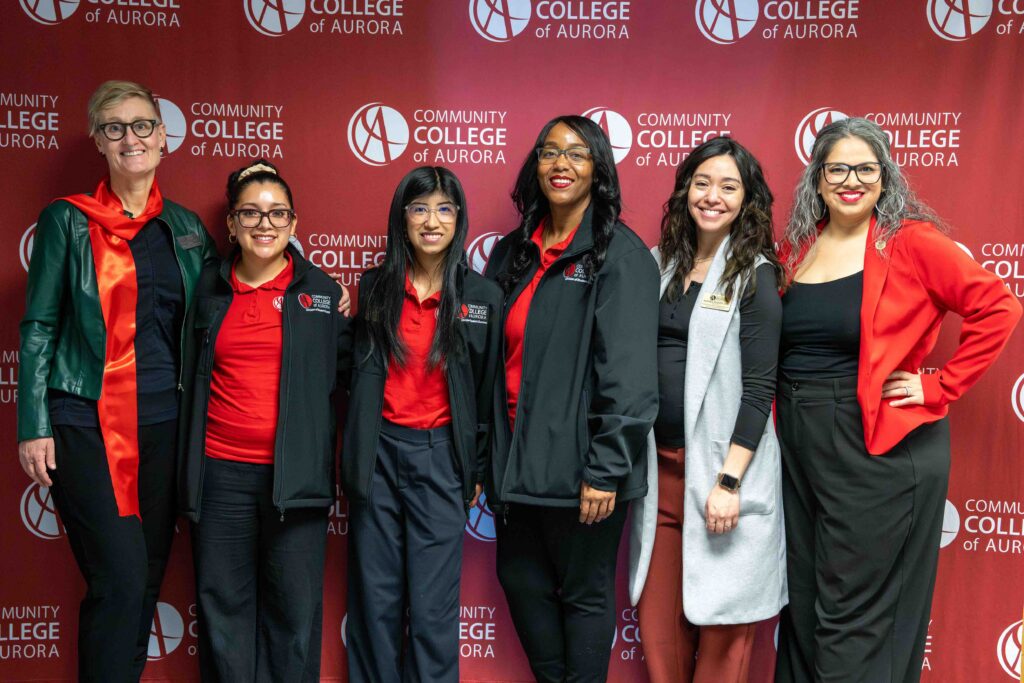 A group of six people stands in front of a “Community College of Aurora” backdrop. They are smiling, dressed in business-casual attire with red accents, exuding a friendly and professional vibe.