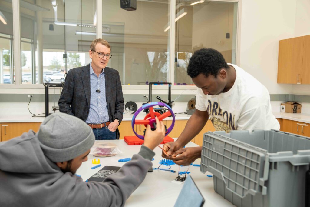 In a bright workshop, two individuals collaborate on a project with colorful gears, observed by a man in a suit.