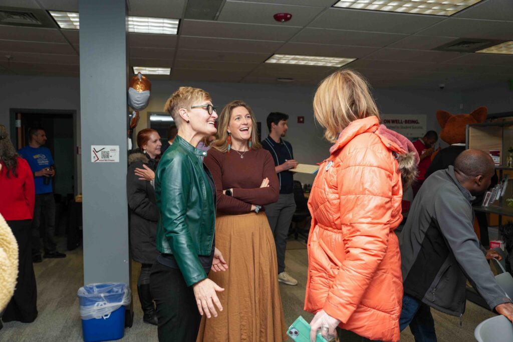 A group of people socialize at an indoor event. Three women in bright, casual attire are engaged in conversation, smiling and laughing. Others mingle in the background, creating a lively and friendly atmosphere.