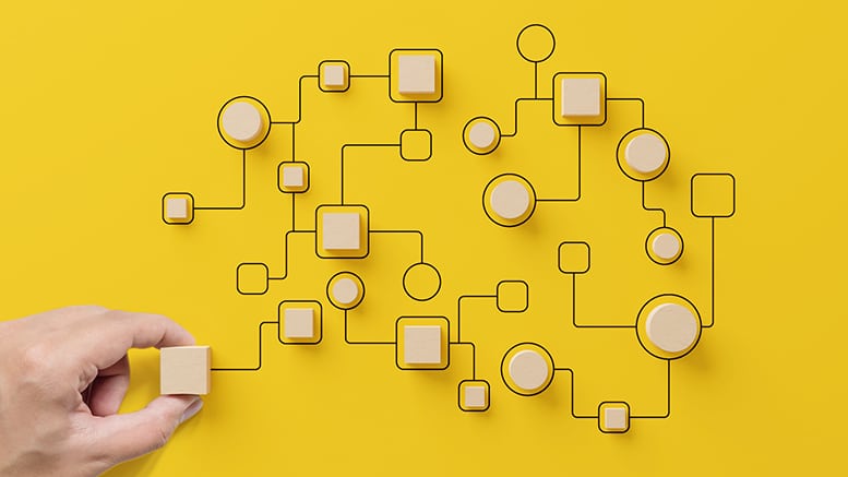 A hand places a wooden block among interconnected circular and square nodes on a bright yellow background, resembling a network or flowchart.