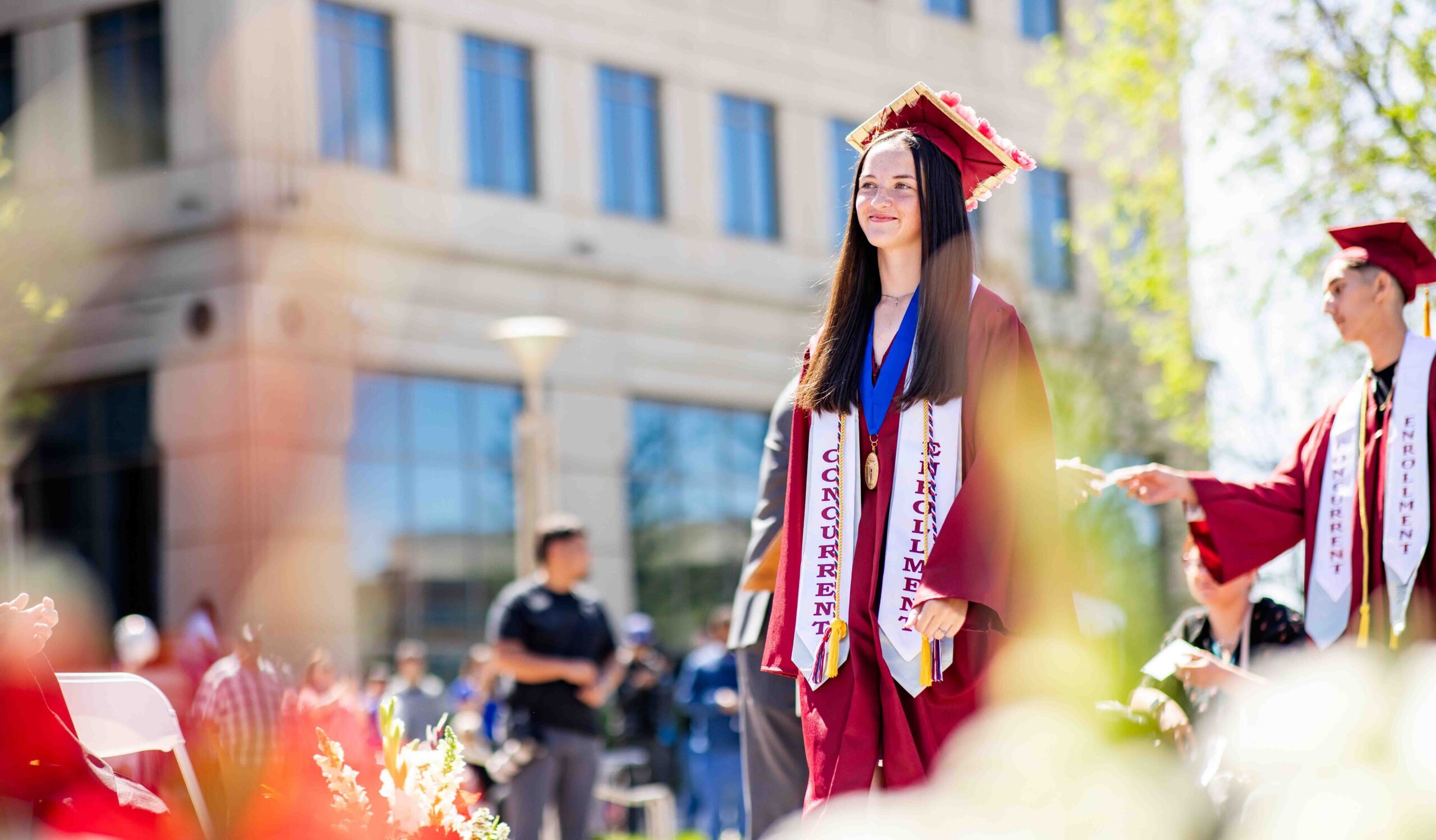 Graduate stands proudly at an outdoor ceremony in crimson cap and gown with honor stoles that read 'Concurrent Enrollment.' Bright sunlight, blurred audience, and flowers create a celebratory mood.