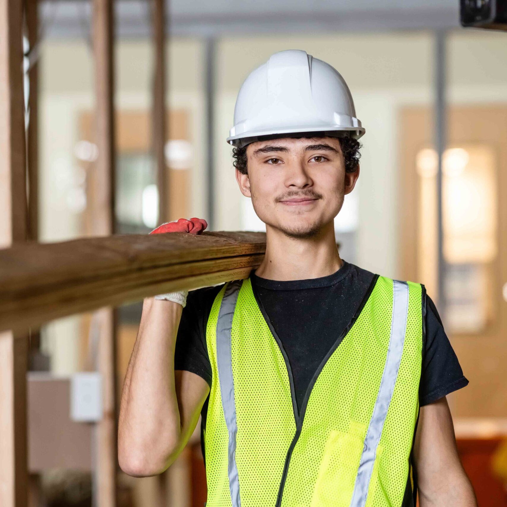 A construction worker in a white hard hat and yellow safety vest carries wooden planks on his shoulder, smiling confidently indoors at a building site.