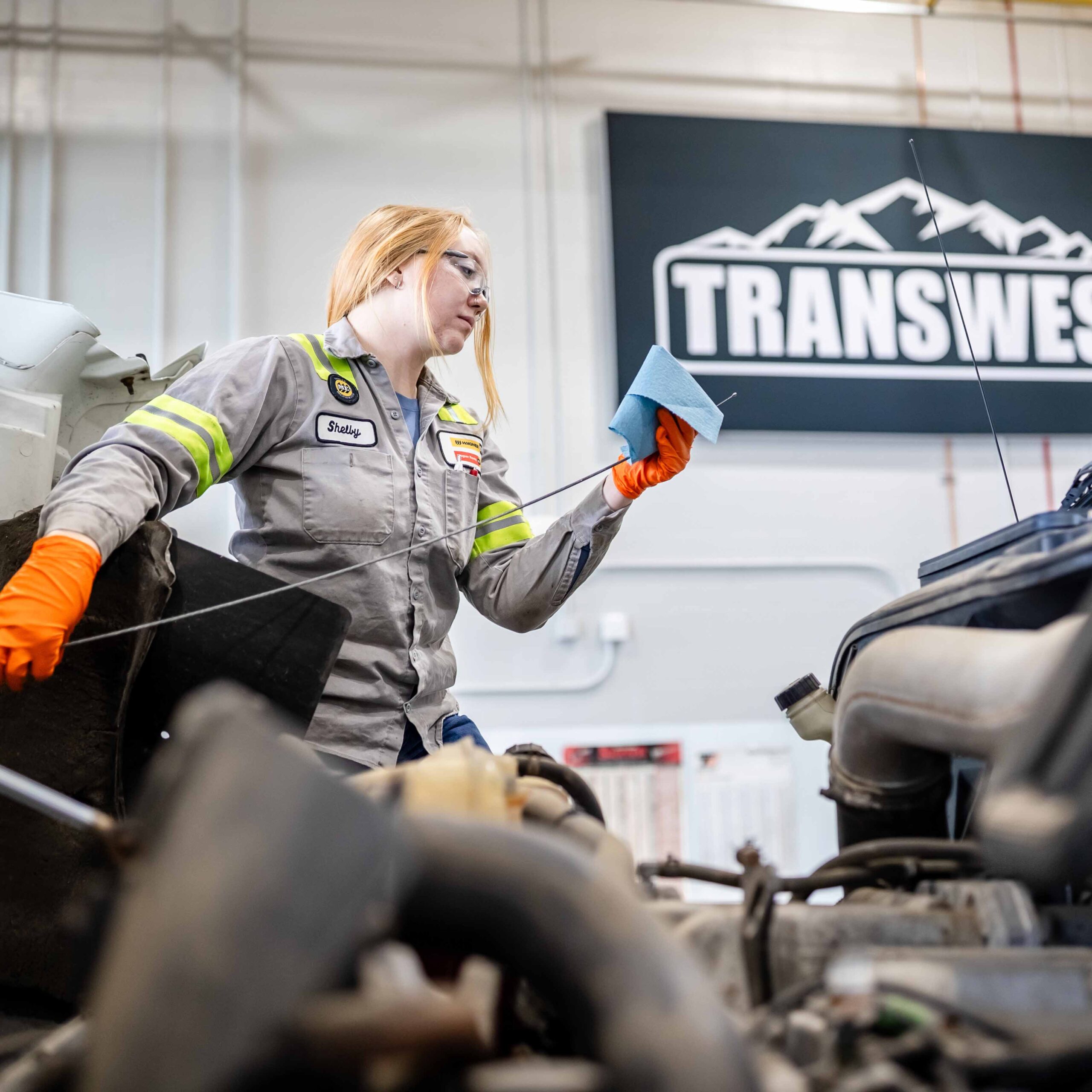 A mechanic wearing orange gloves and a gray uniform checks an engine with a dipstick and blue cloth in a workshop.