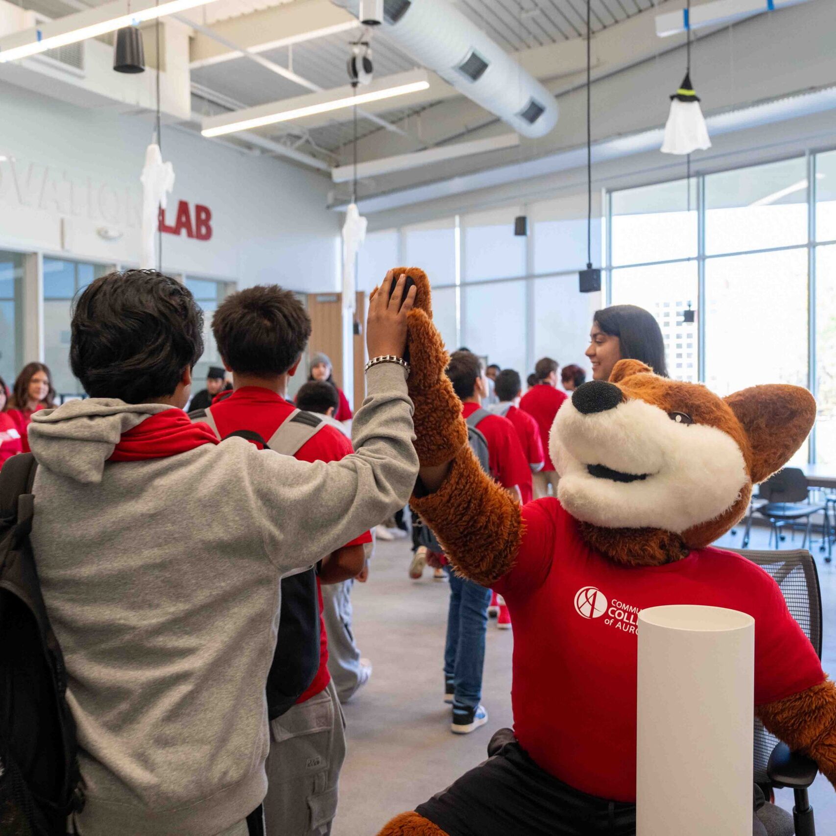 A child in a gray hoodie gives a high five to a fox mascot in a red shirt in a lively room labeled 