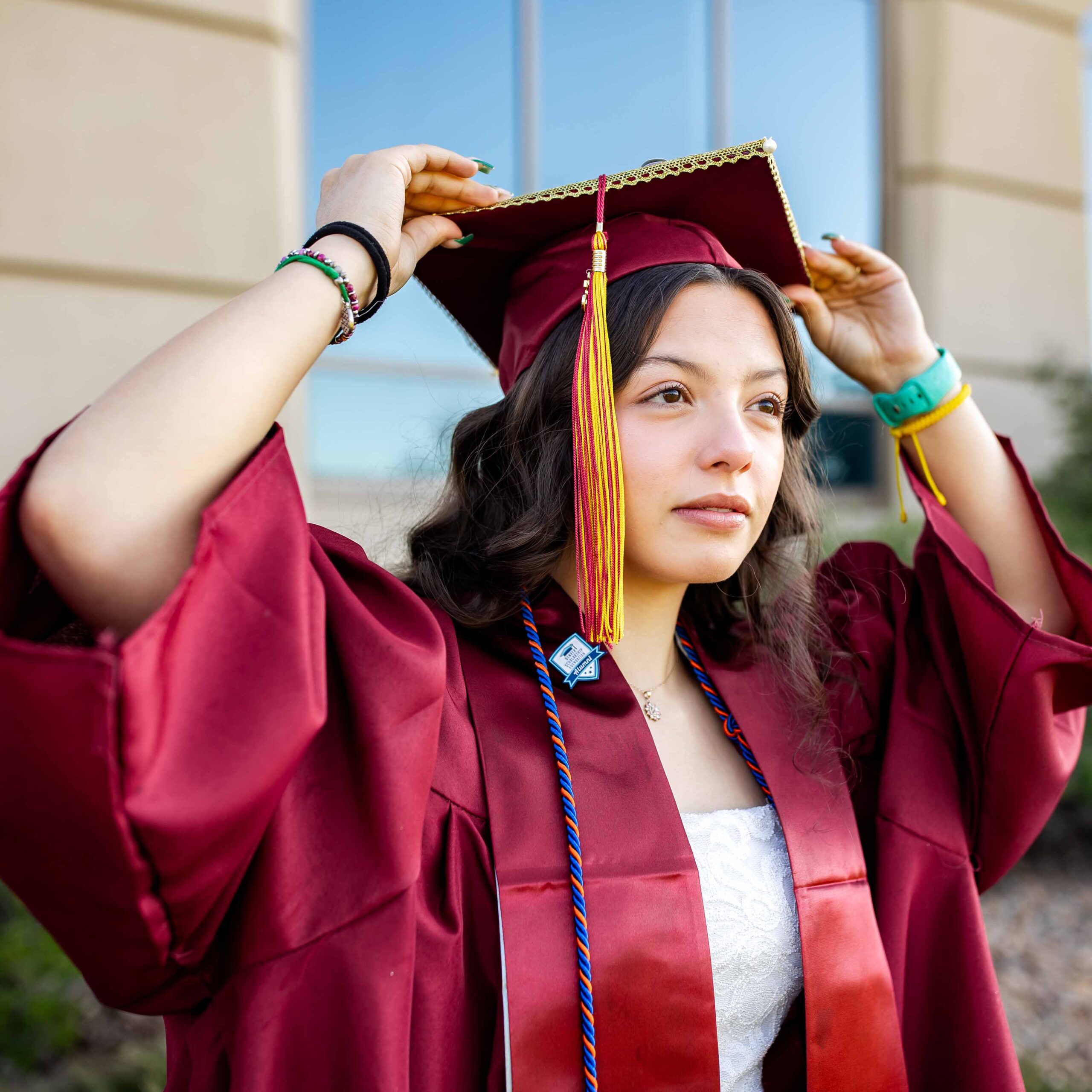 Young woman in maroon graduation cap and gown adjusts her cap, looking confidently ahead. Background shows a building exterior in soft focus.