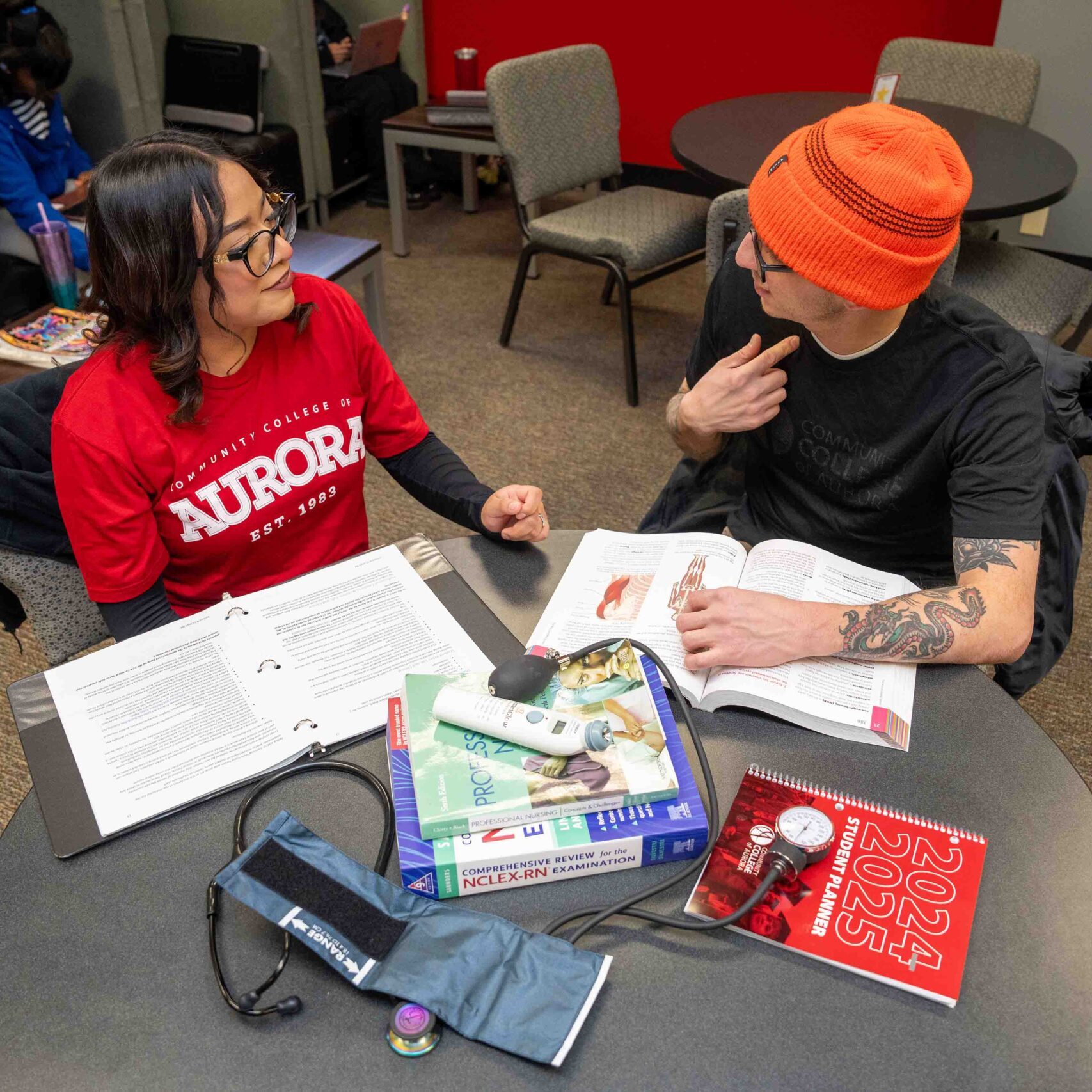 Two people collaborate at a round table with textbooks and medical equipment. One wears a red shirt, the other an orange beanie, creating a focused atmosphere.