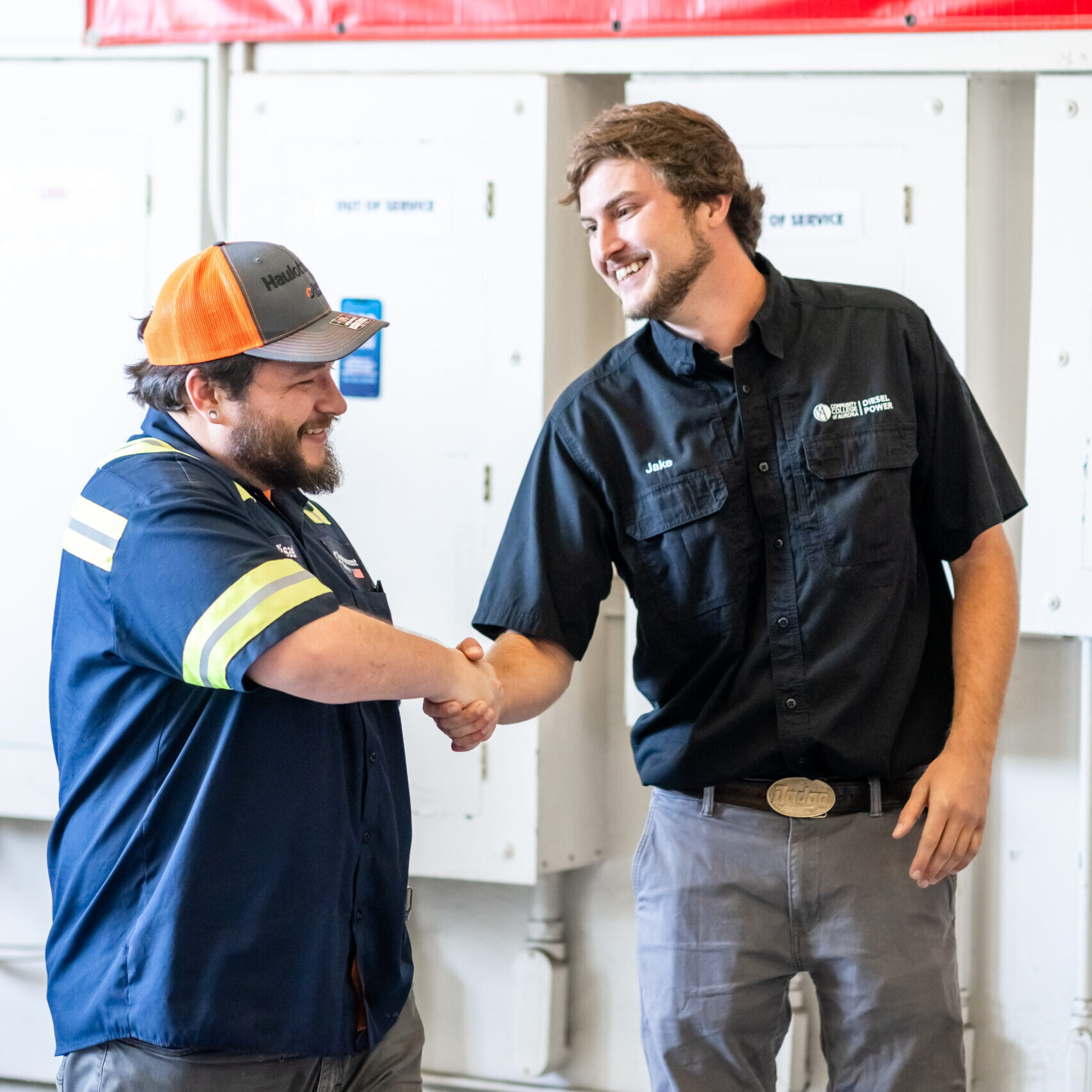 Two men in work uniforms shake hands, smiling warmly. They're standing in front of panels labeled 