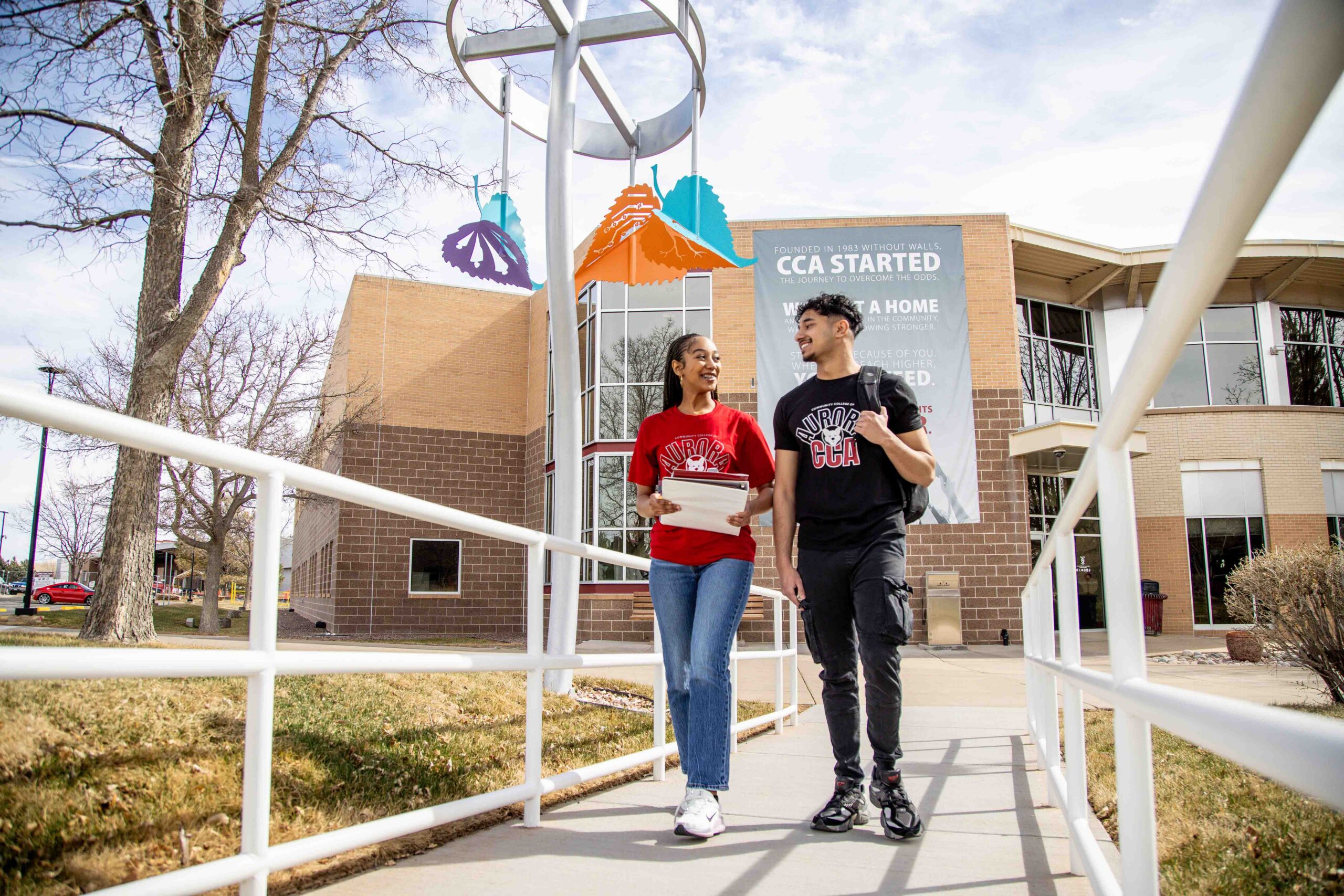 Two students walk along a path, smiling and talking. They're in front of a brick building. The atmosphere is casual and sunny.