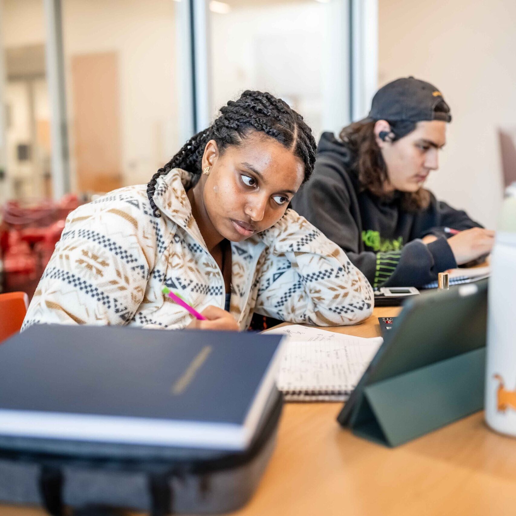 Two students study at a table, one writing in a notebook, appearing focused. Laptops, books, and a water bottle are visible. The setting is a bright, modern room.